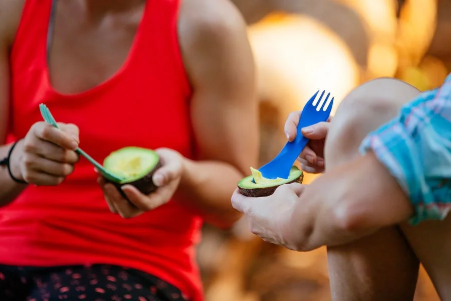 Two people sitting outdoors, each holding half of an avocado and using sporks to eat the fruit.