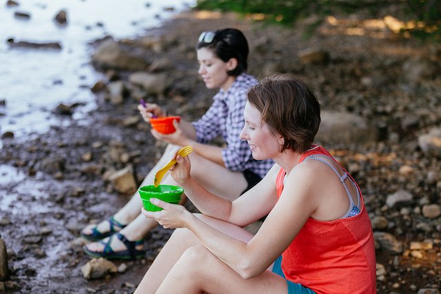 Two people sitting on a rocky shore eating from bowls with spoons.