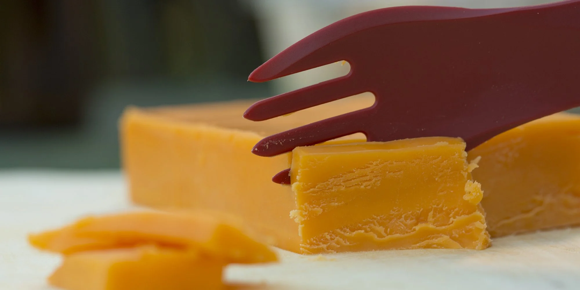 Close-up of a block of cheddar cheese being sliced with a red cheese slicer on a cutting board.