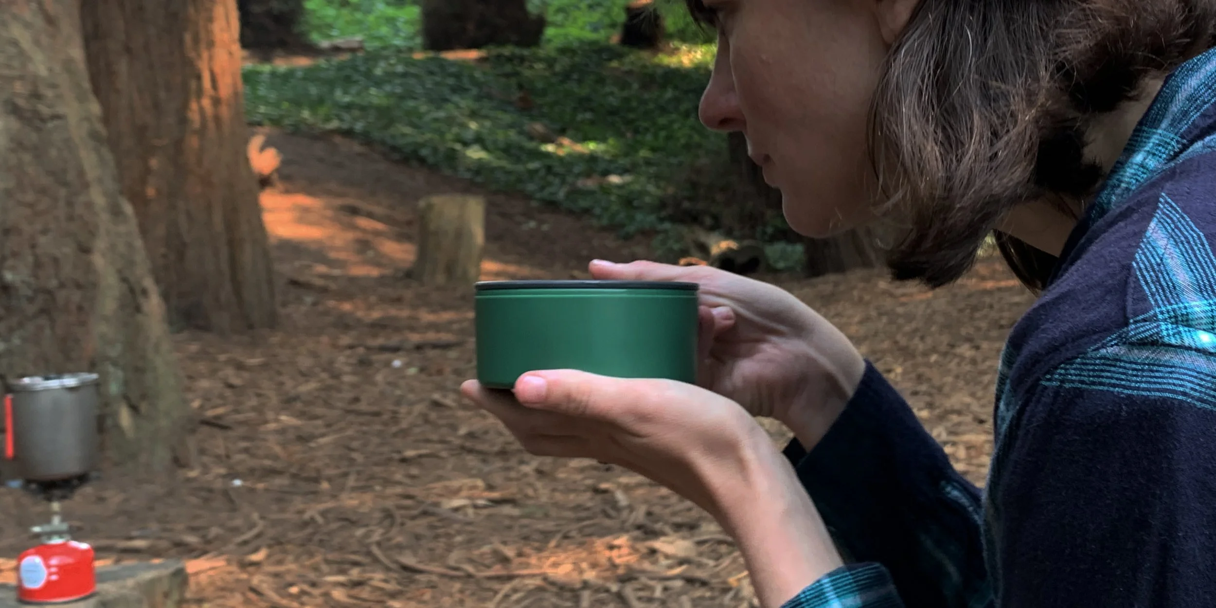 Person holding a green cup in a forest with a camping stove on a tree stump.