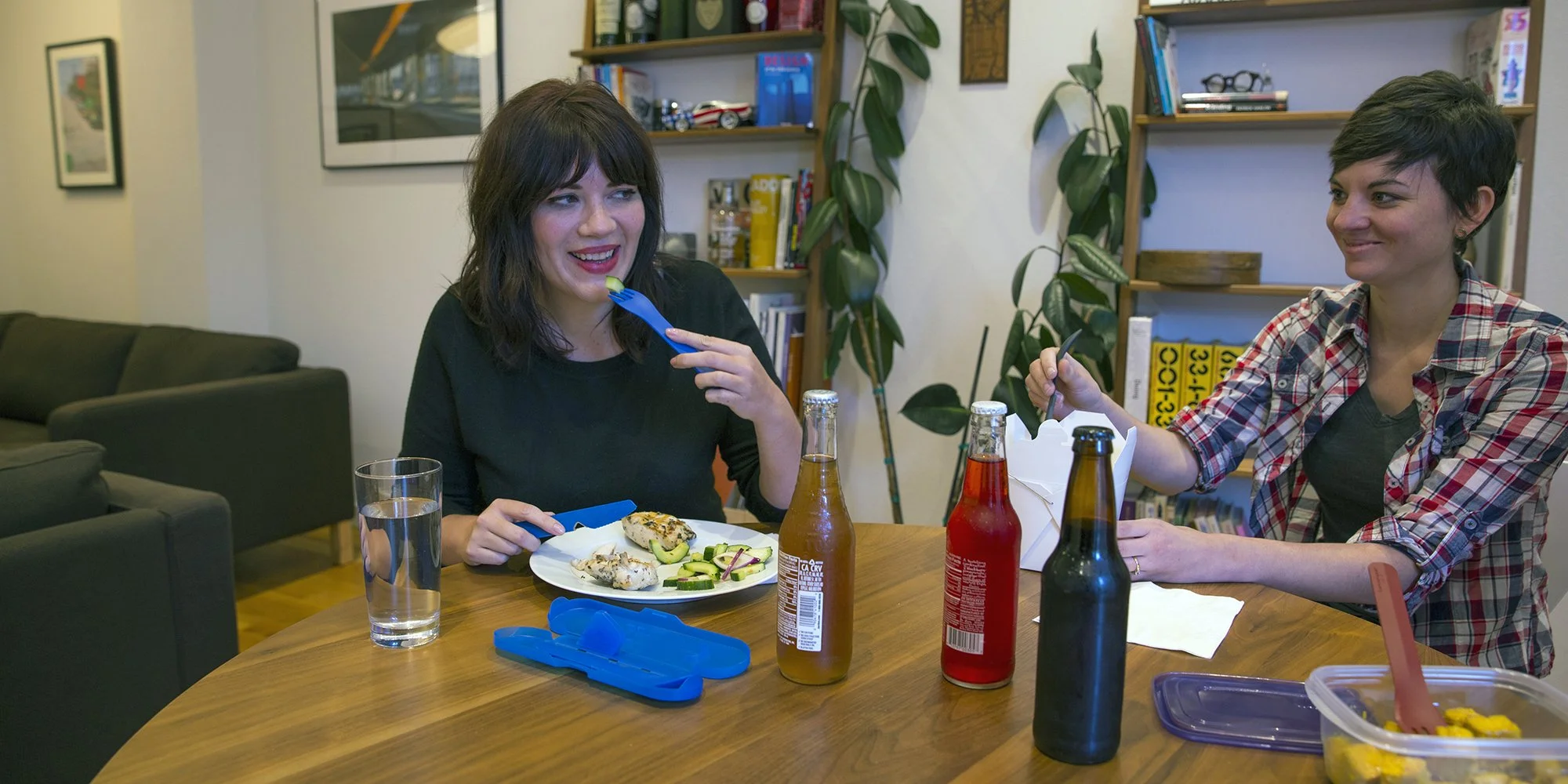 Two women sitting at a dining table eating and smiling, with plates of food, beverages in bottles, and a glass of water. The room has bookshelves and a couch in the background.