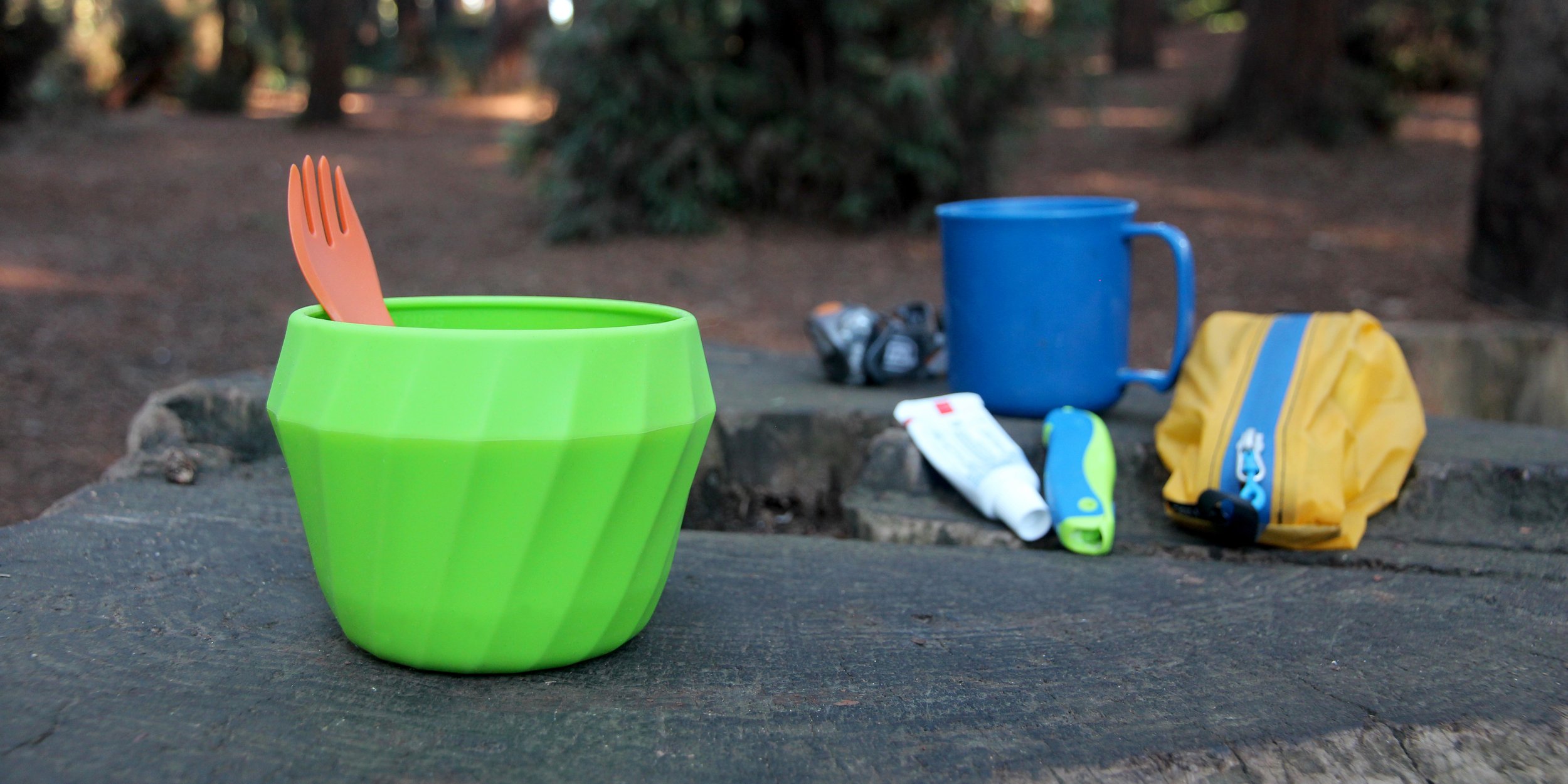 Camping gear on wooden surface, featuring a green bowl with orange spork, blue mug, and yellow zip pouch, in a forest setting.