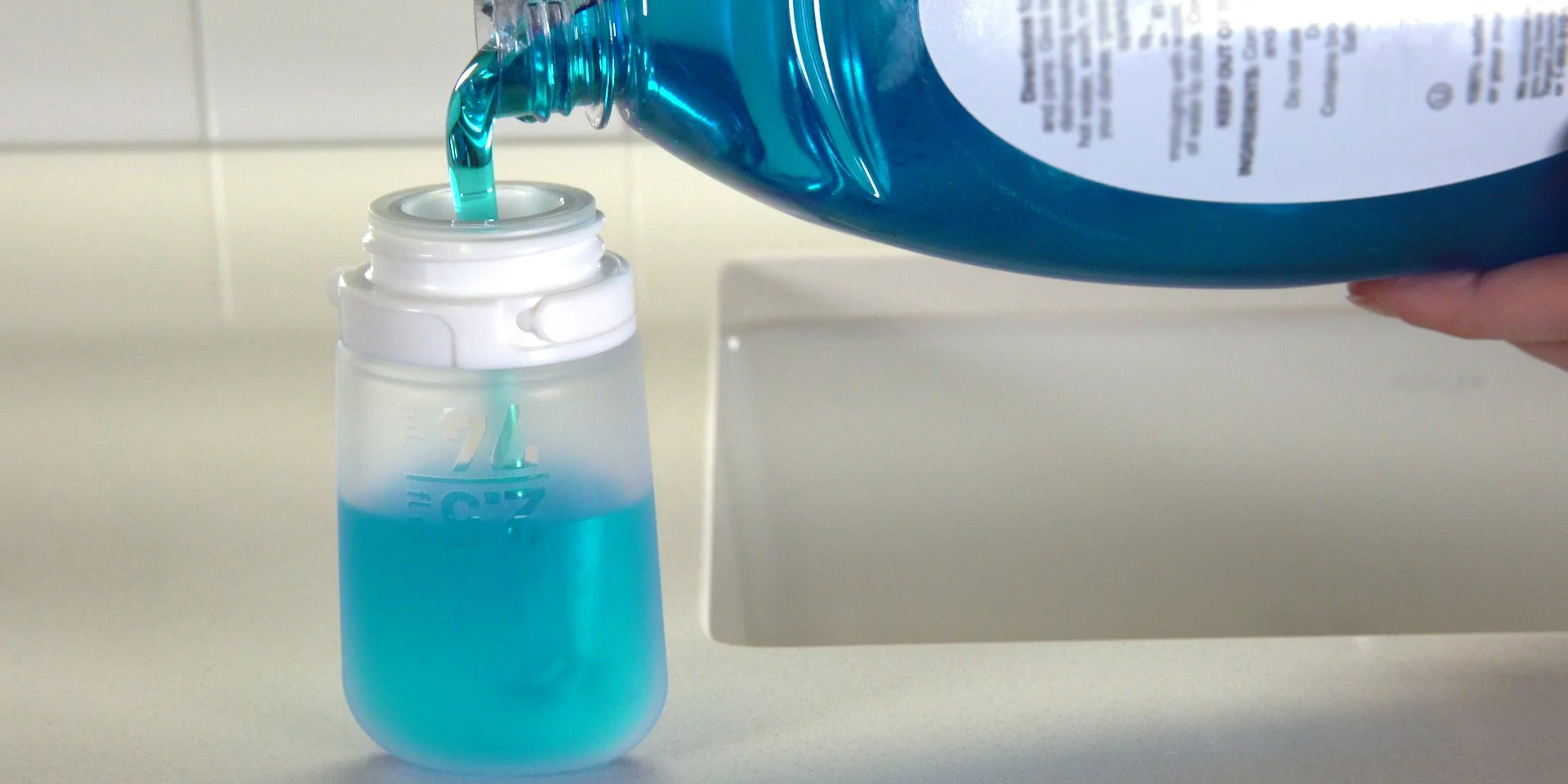 Pouring blue mouthwash into a clear container on a countertop.