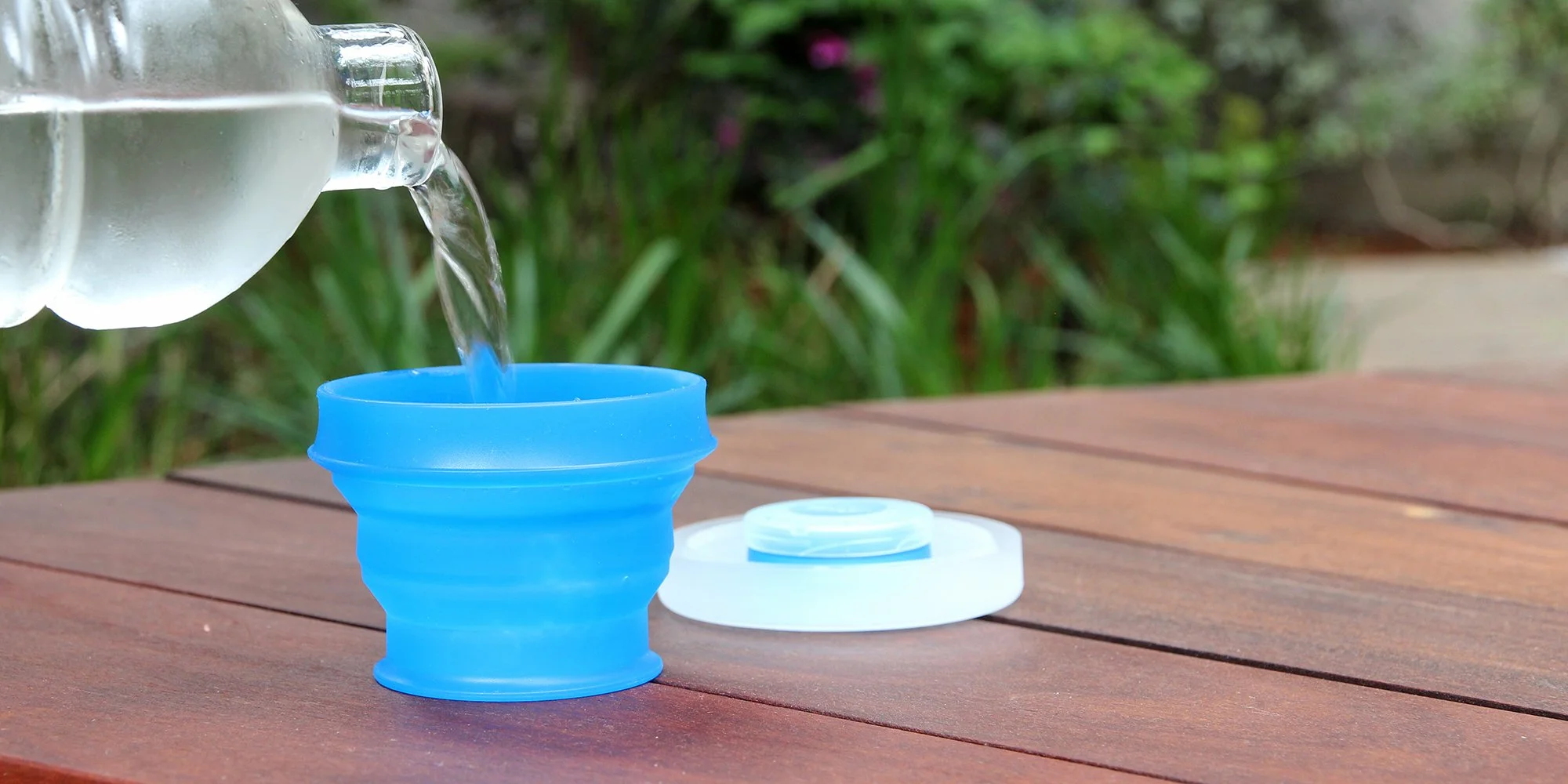 Water being poured from a bottle into a blue collapsible cup on a wooden surface with a lid nearby.