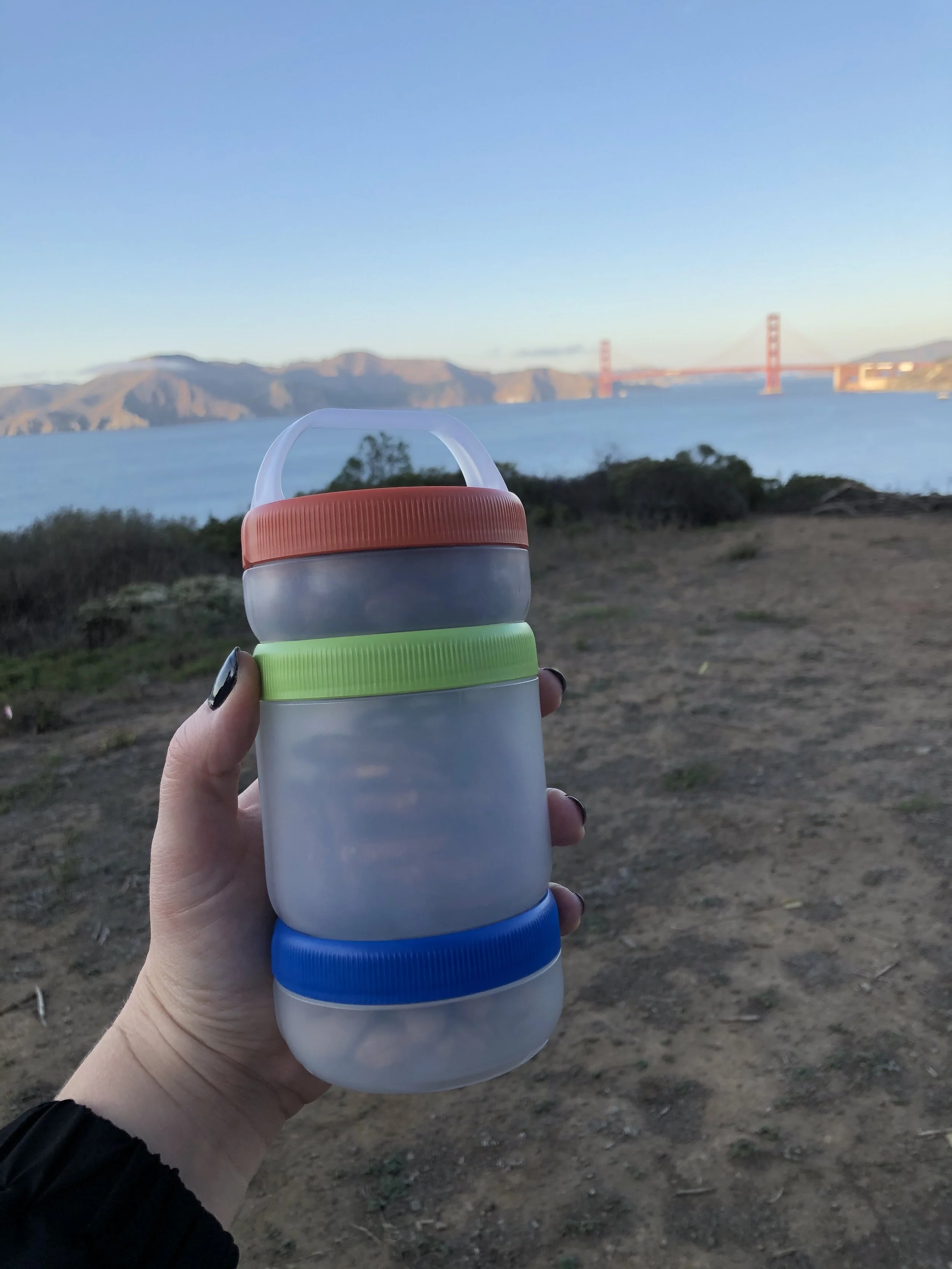 Hand holding a stackable container with colorful lids in front of a scenic landscape with a bridge and mountains.