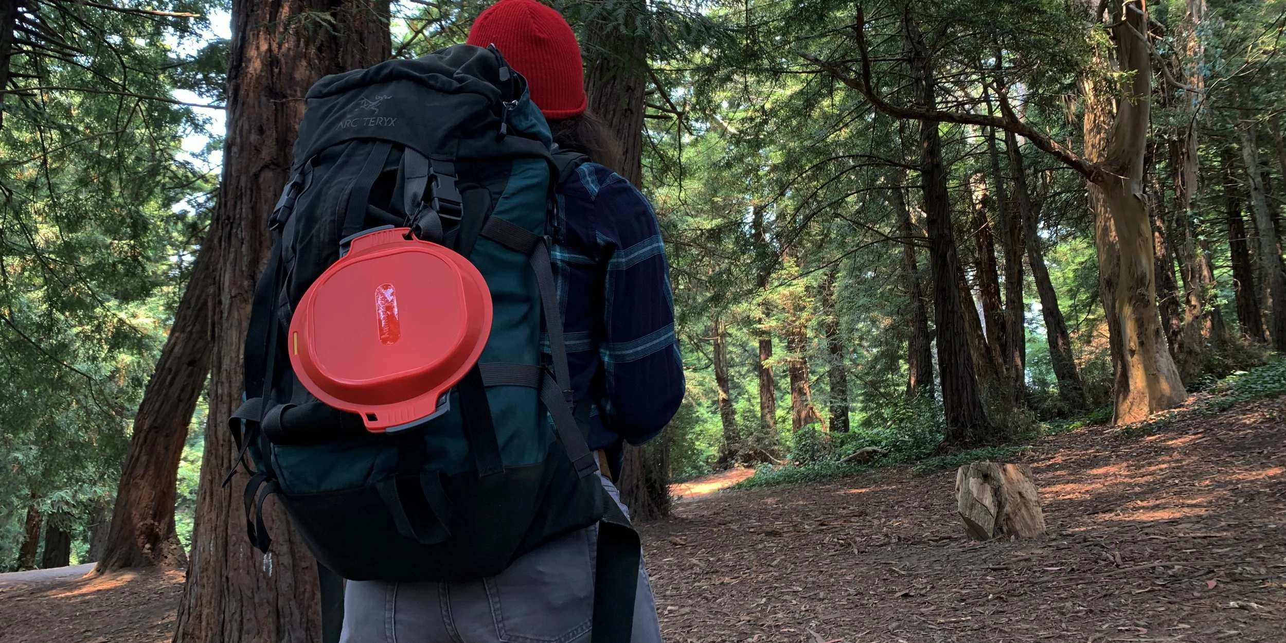 Person with a large backpack and a red case hiking in a wooded area.