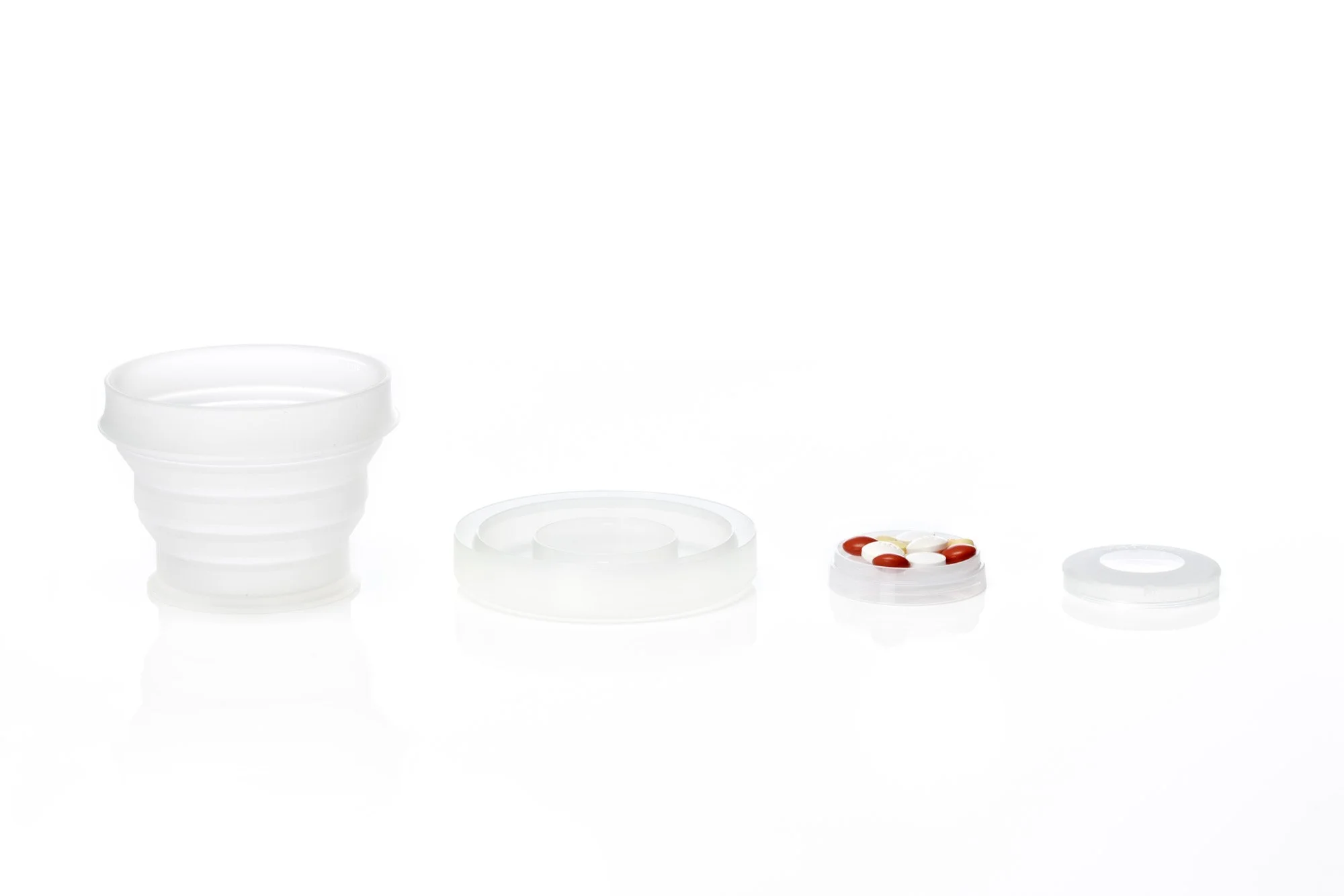 Collapsible silicone cup with lid, storage container, and white and red pills on a white surface.