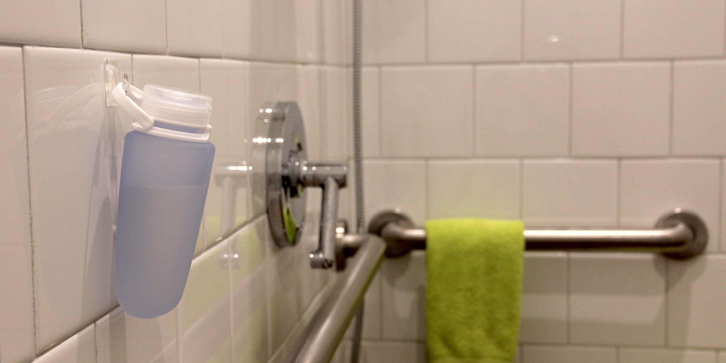 Shower area with white tiled walls, a suction cup hanging a silicone travel bottle, a metallic faucet and handle, and a green towel on a metal towel bar.
