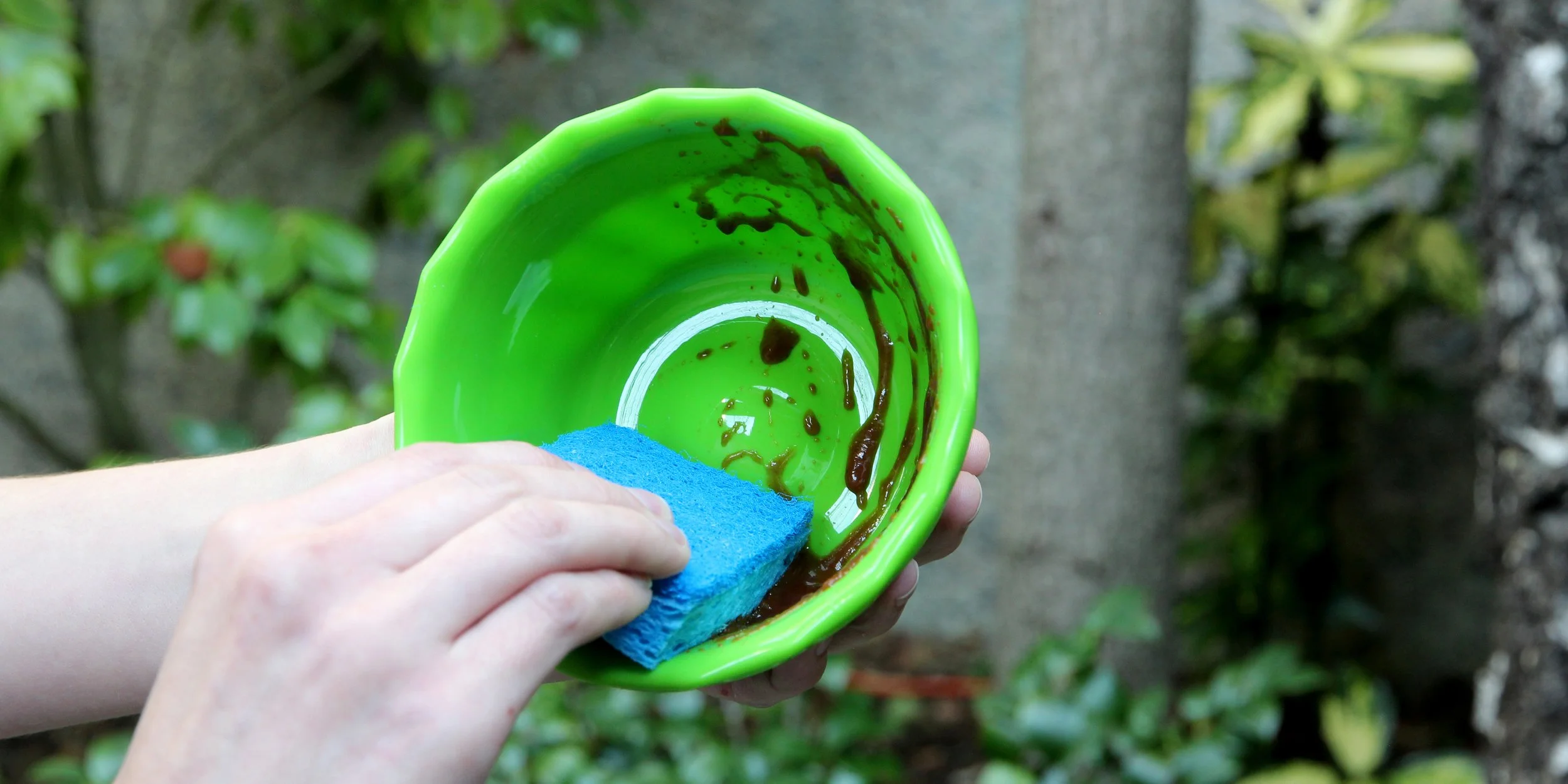 Person cleaning a green bowl with a blue sponge outdoors.