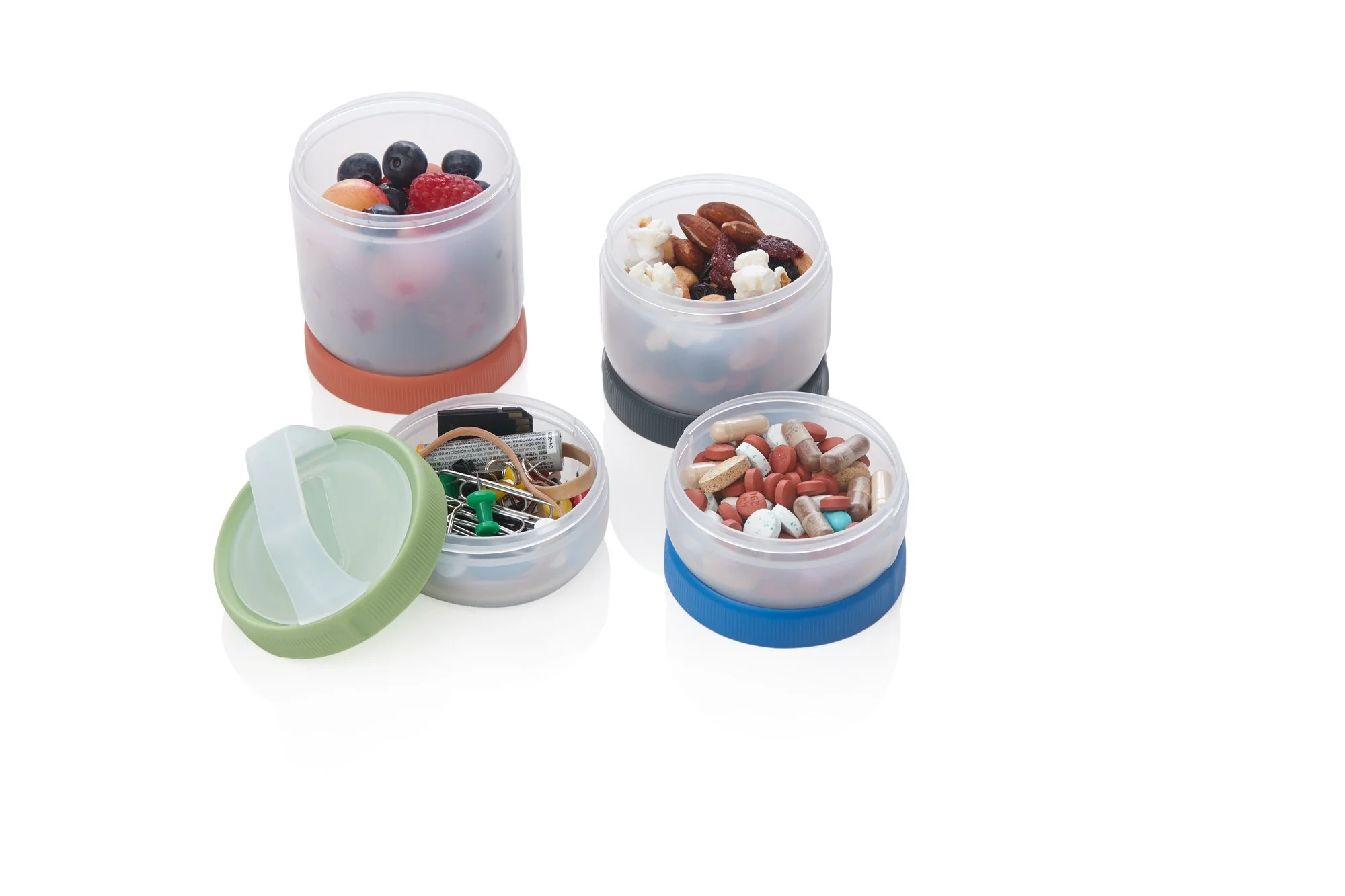 Four small open containers with various contents: one with mixed berries, one with nuts and popcorn, one with office supplies like paperclips and pushpins, and one with assorted pills and capsules, all on white background.