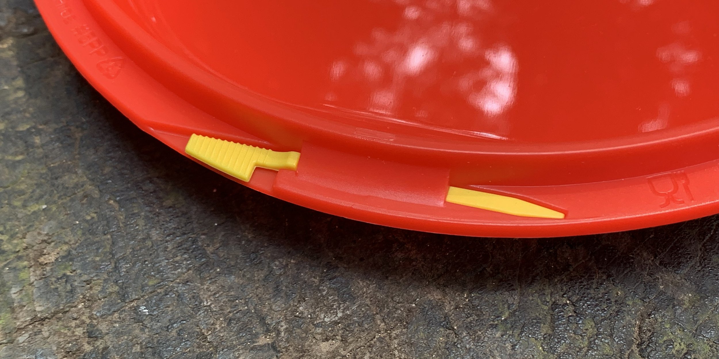 Close-up of a red plastic lid with a yellow pull tab, placed on a textured surface.