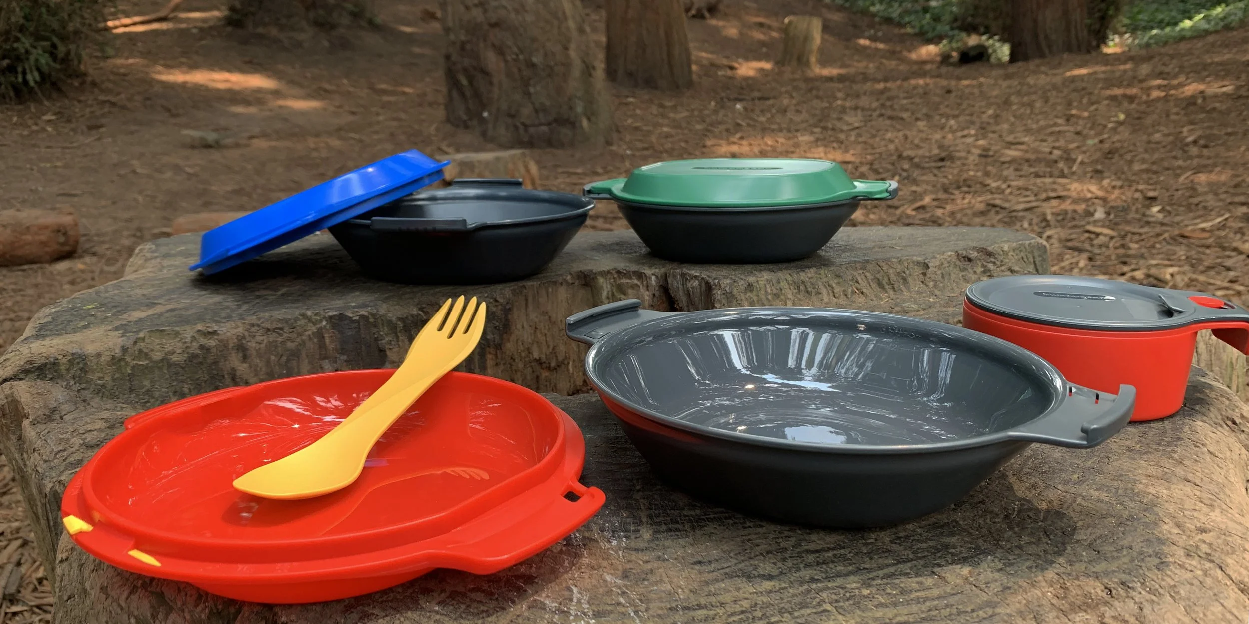 Camping cookware and utensils on a tree stump in the woods, including a yellow spork, red and blue lids, and gray and red bowls and pots.