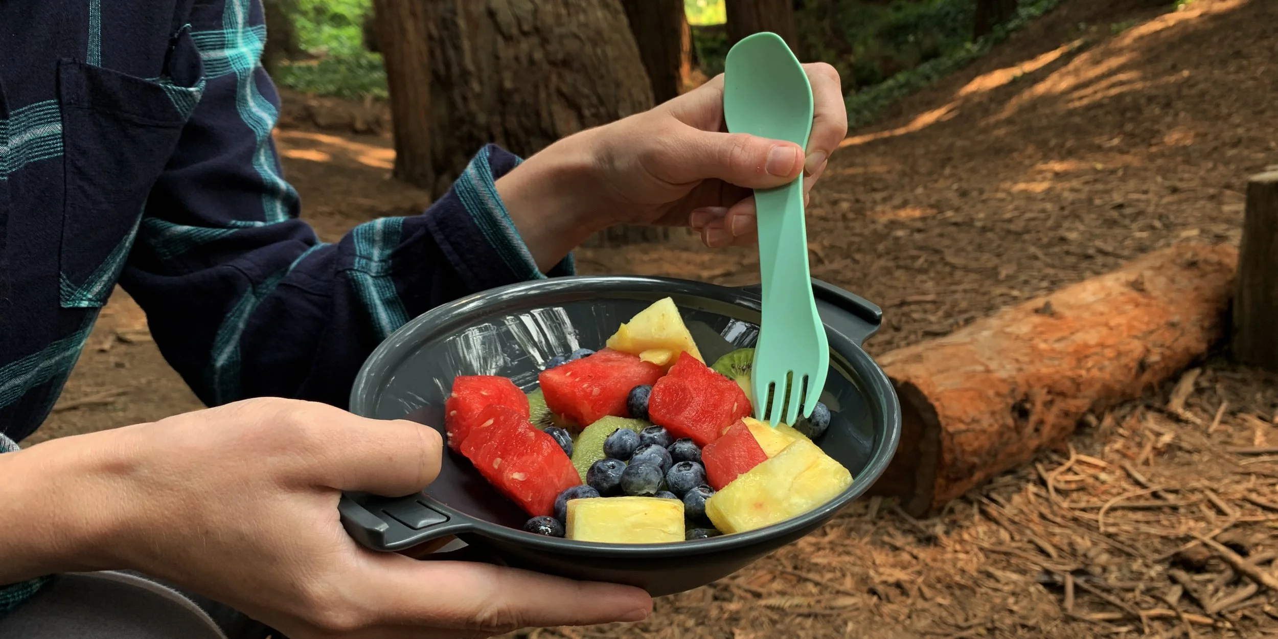 Person holding a bowl of fruit salad with watermelon, pineapple, blueberries, and kiwi using a teal spork in a forest setting.