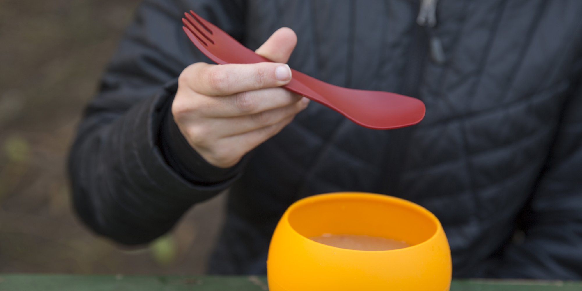 Person holding a red spork above an orange bowl outdoors.