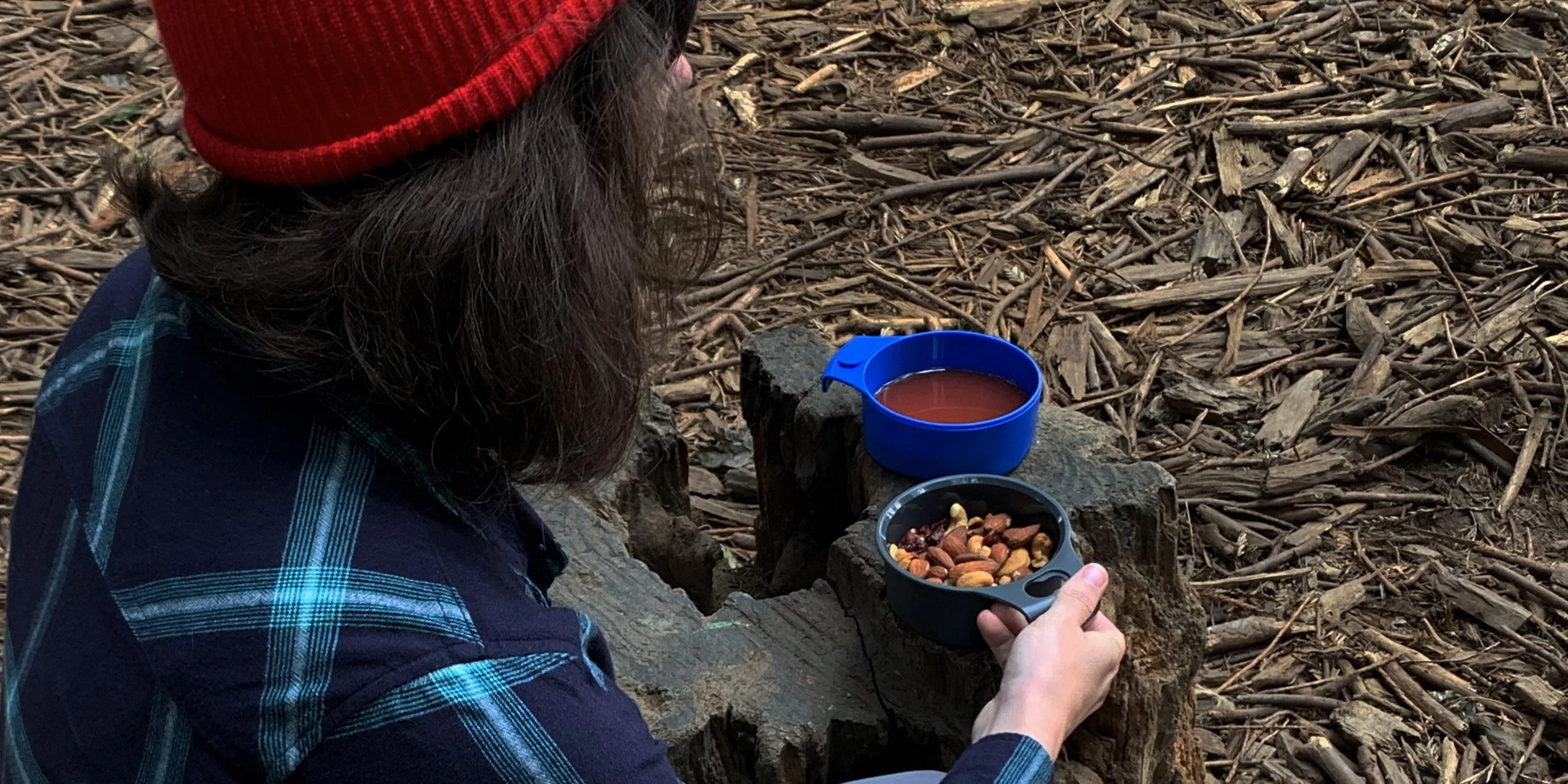 Person wearing a red beanie and plaid shirt sitting in the woods, holding a bowl of mixed nuts, with a cup of red liquid on a tree stump.