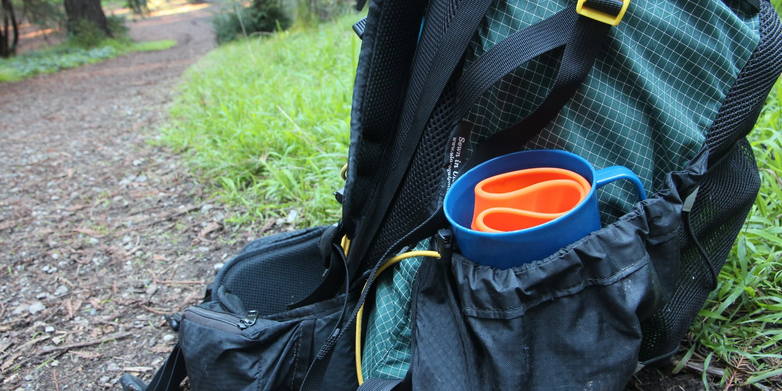 Backpack with a blue and orange mug in side pocket, on a forest trail.