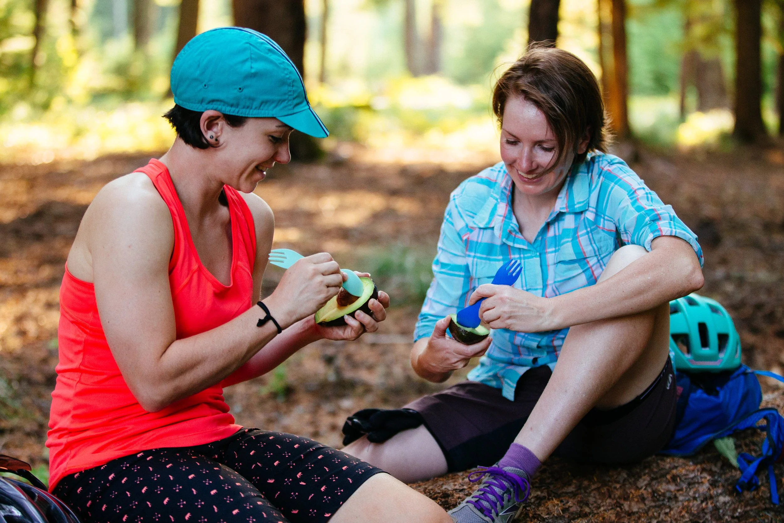 Two people sitting in a forest setting, eating avocados with forks.