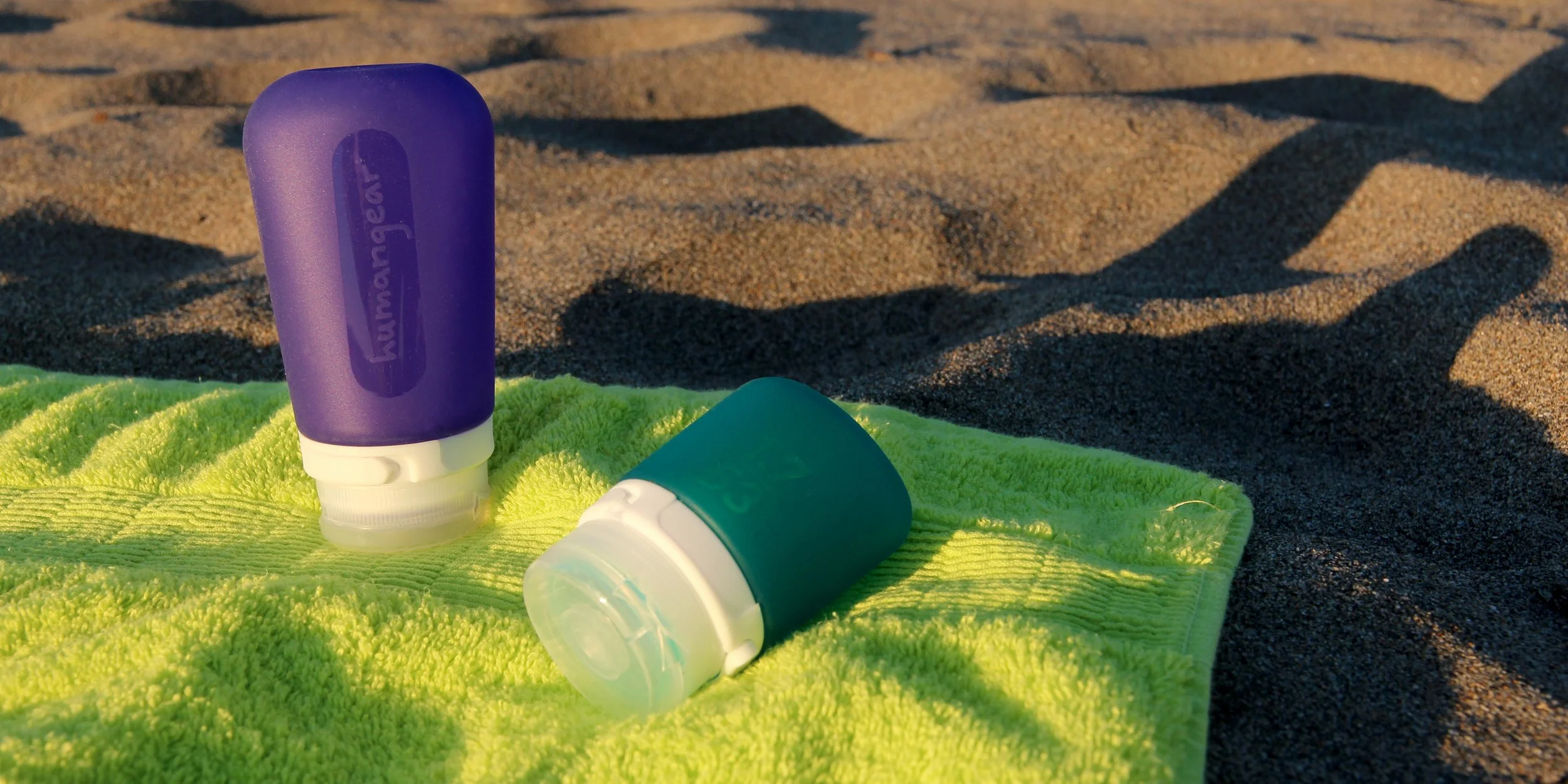 Two travel-sized toiletry bottles on a green towel at the beach, with sand in the background.
