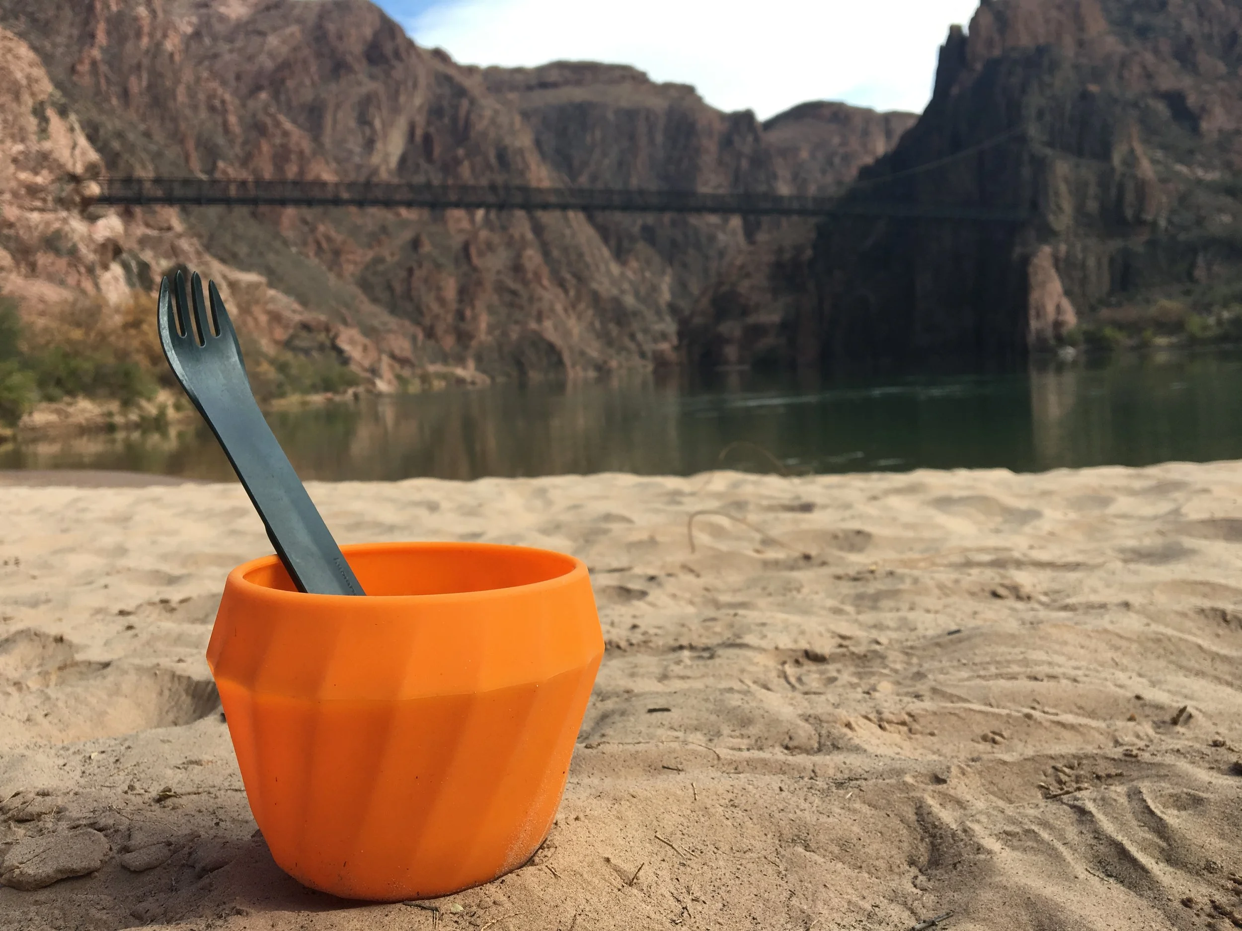 Orange cup with a spork on sandy riverbank, bridge and rocky cliffs in the background.
