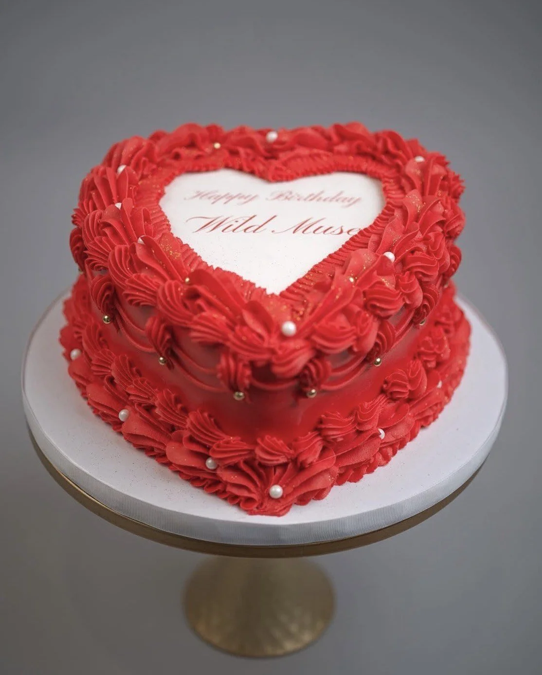 A red heart-shaped cake decorated with vintage-style buttercream piping, edible pearls, gold accents, and a personalized message in the center, displayed on a white board and gold cake stand.
