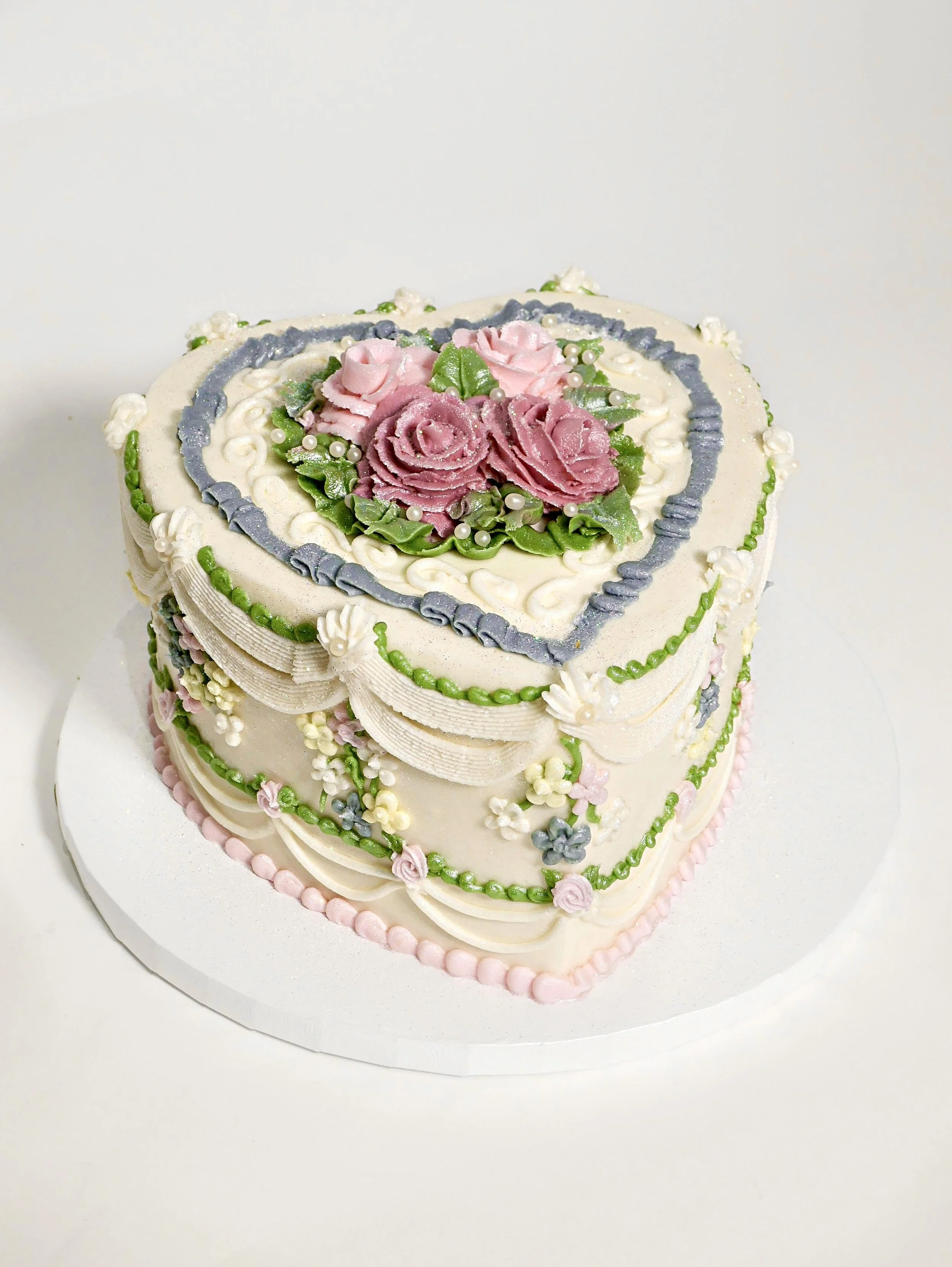 Heart-shaped vintage-style cake decorated with piped buttercream swags, green detailing, and pink and purple roses in the center, set on a white cake board.