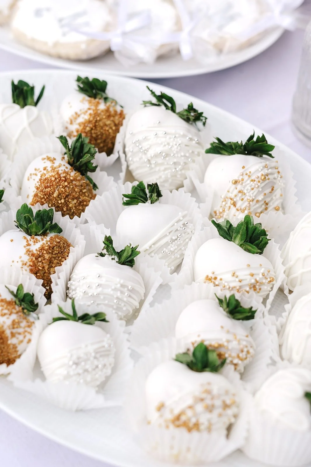 White chocolate–covered strawberries decorated with gold sugar crystals, pearl sprinkles, and white drizzle, arranged in paper cups on a serving platter.