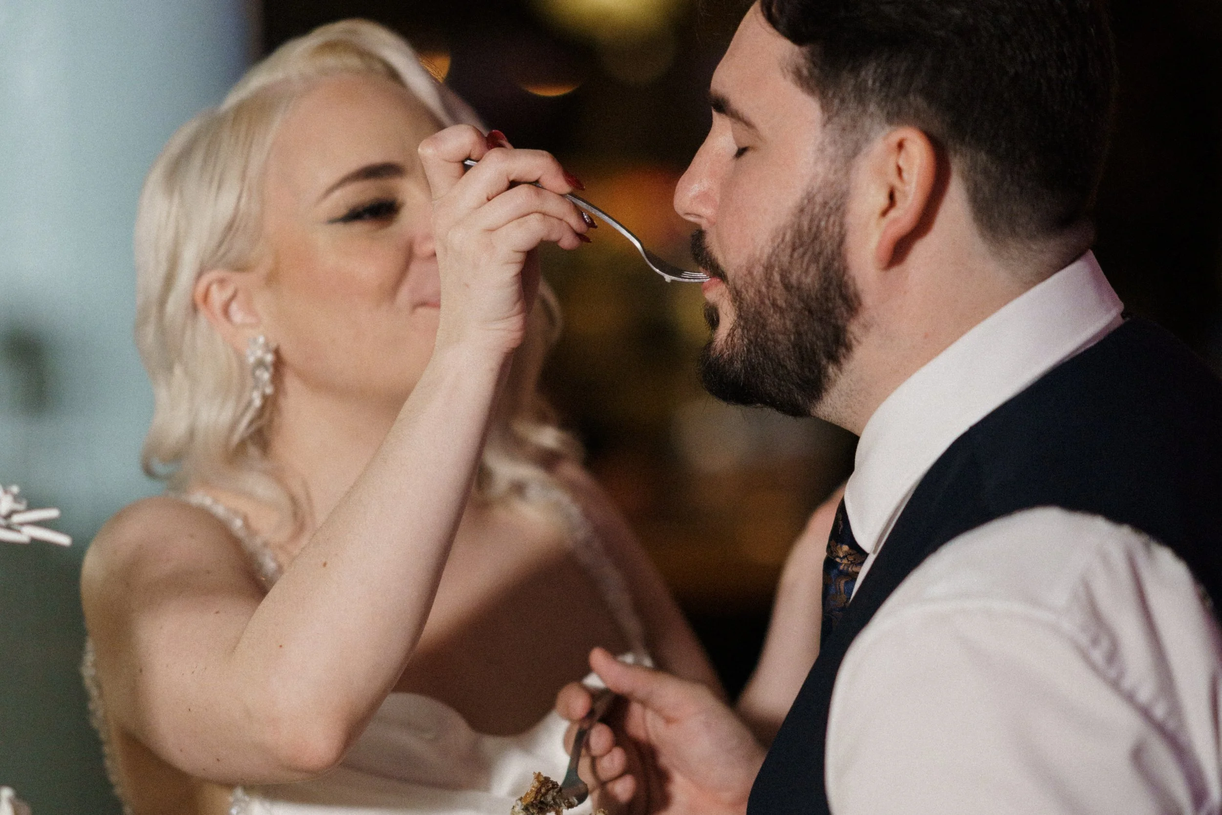 Bride in a white wedding dress playfully feeds a bite of cake to the groom in a vest and tie during their wedding reception.