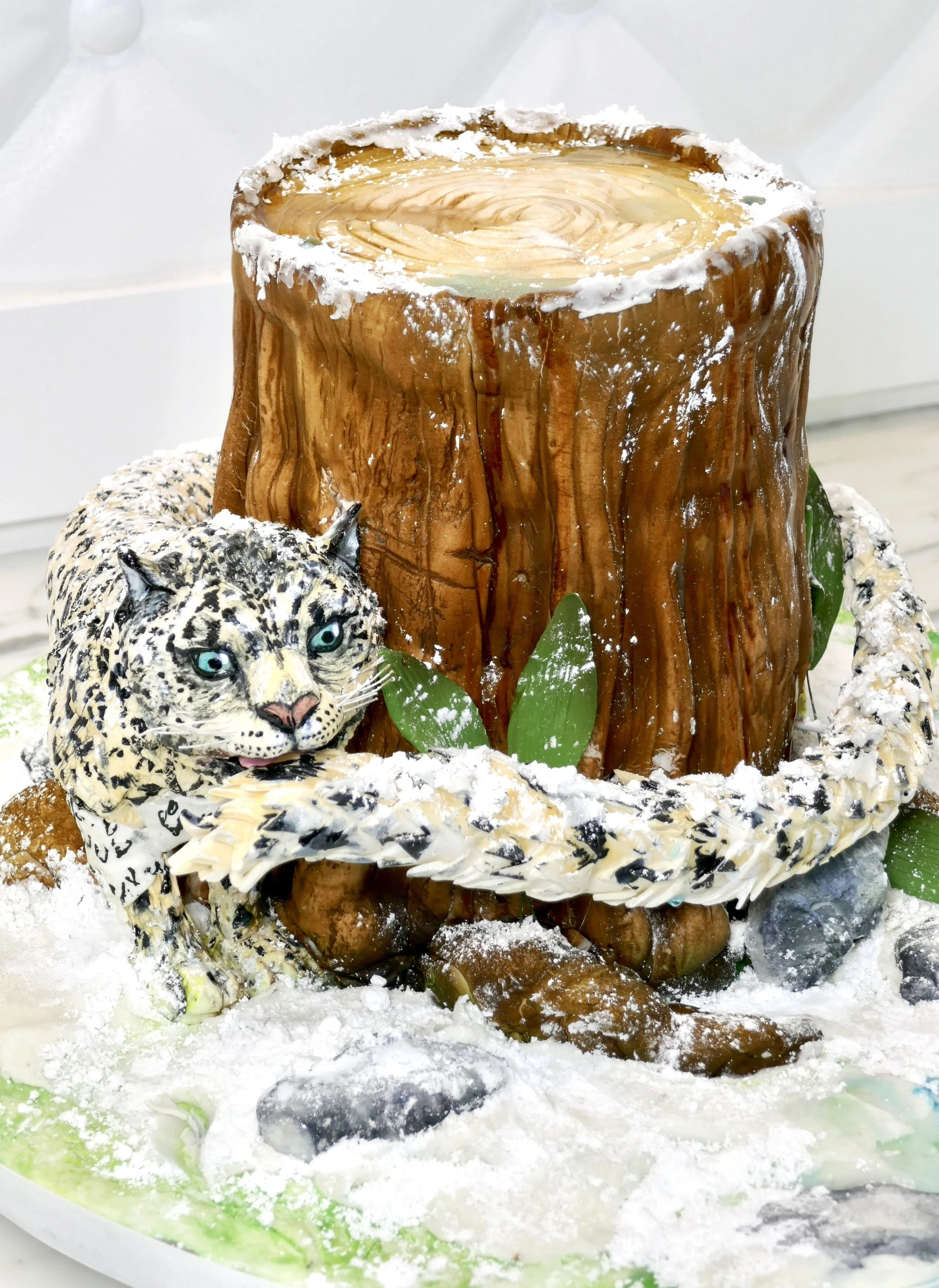 Sculpted 3D cake designed as a snow-covered tree stump with a realistic snow leopard wrapped around the base, featuring detailed fur texture, green leaves, rocks, and powdered sugar to resemble snow.