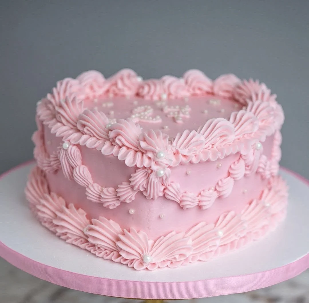 Round pink buttercream cake decorated with piped vintage-style borders, pearl details, and scalloped edges, displayed on a white and pink cake stand.
