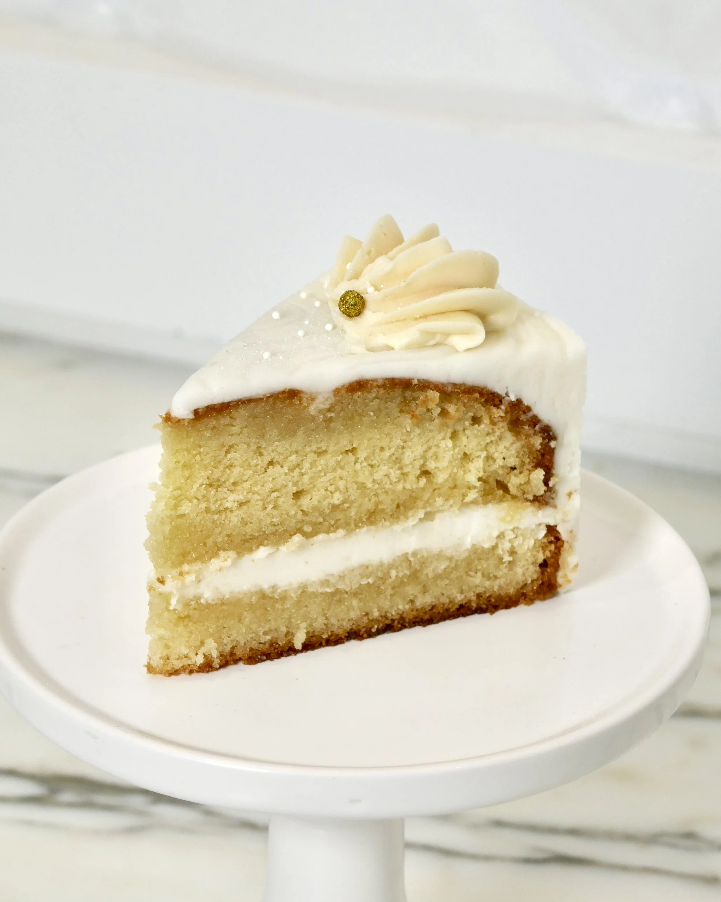 Slice of vanilla cake with a layer of white buttercream filling, frosted in smooth white icing and topped with a piped swirl and gold sprinkle, displayed on a white cake stand.