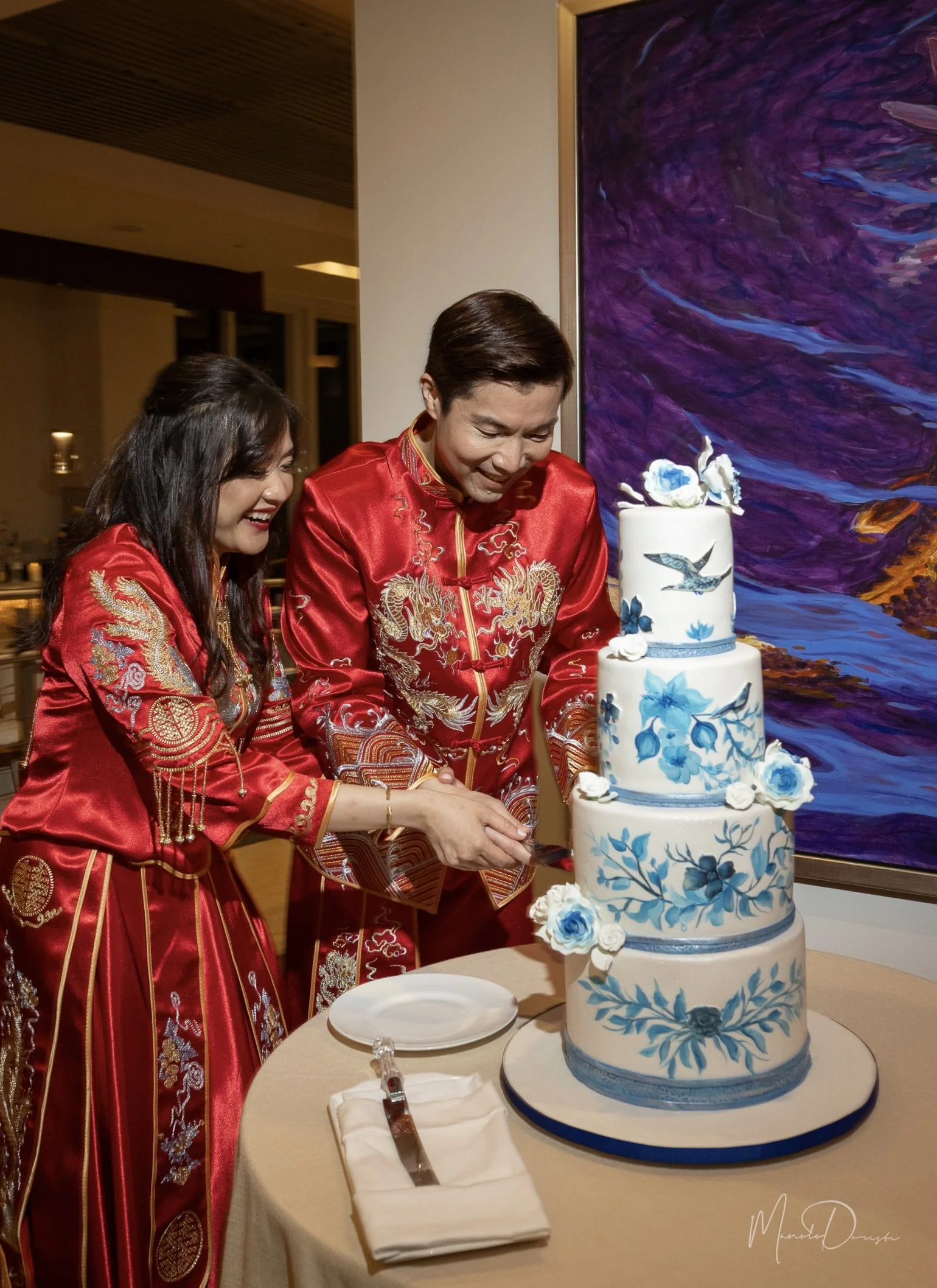 Couple wearing traditional red embroidered attire cut a four-tier white wedding cake decorated with blue floral designs and sugar flowers, standing beside a colorful abstract painting.