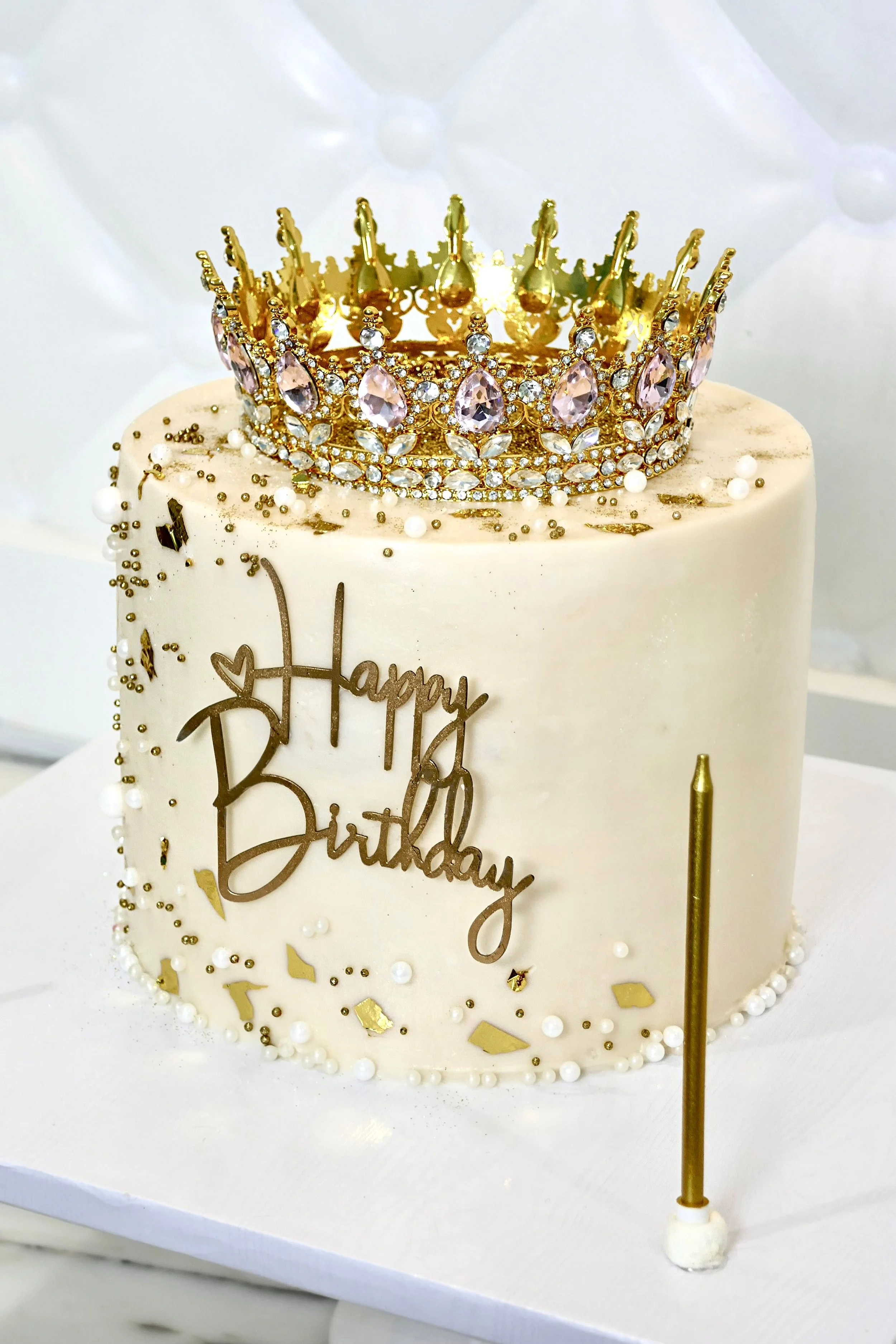 Elegant white birthday cake decorated with gold leaf, pearl accents, and a jeweled gold crown on top, featuring a “Happy Birthday” topper and a tall gold candle beside it.