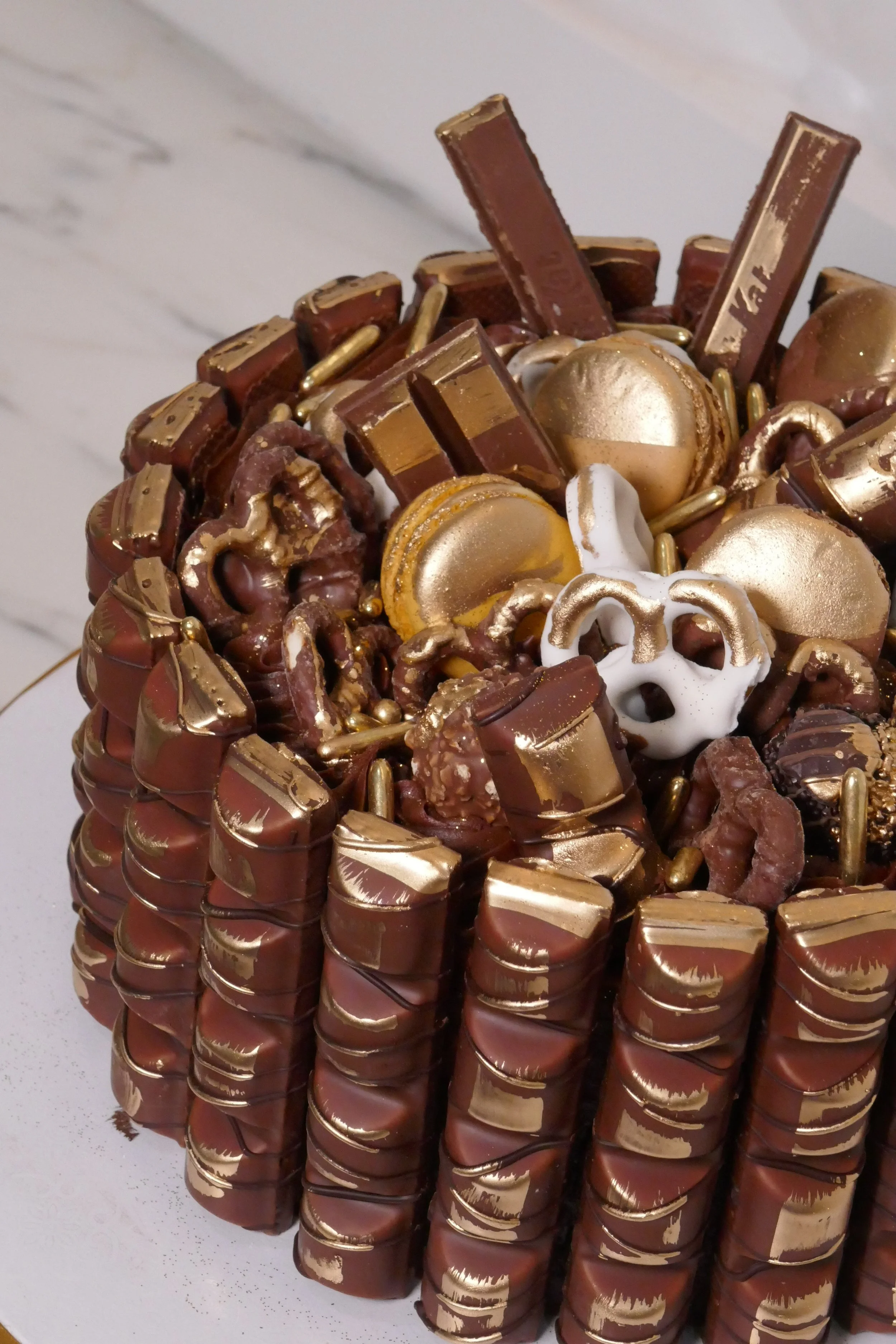 Zoomed in image of round chocolate celebration cake wrapped with rows of chocolate bars and topped with gold-dusted macarons, pretzels, and truffles, displayed on a white and gold cake stand.