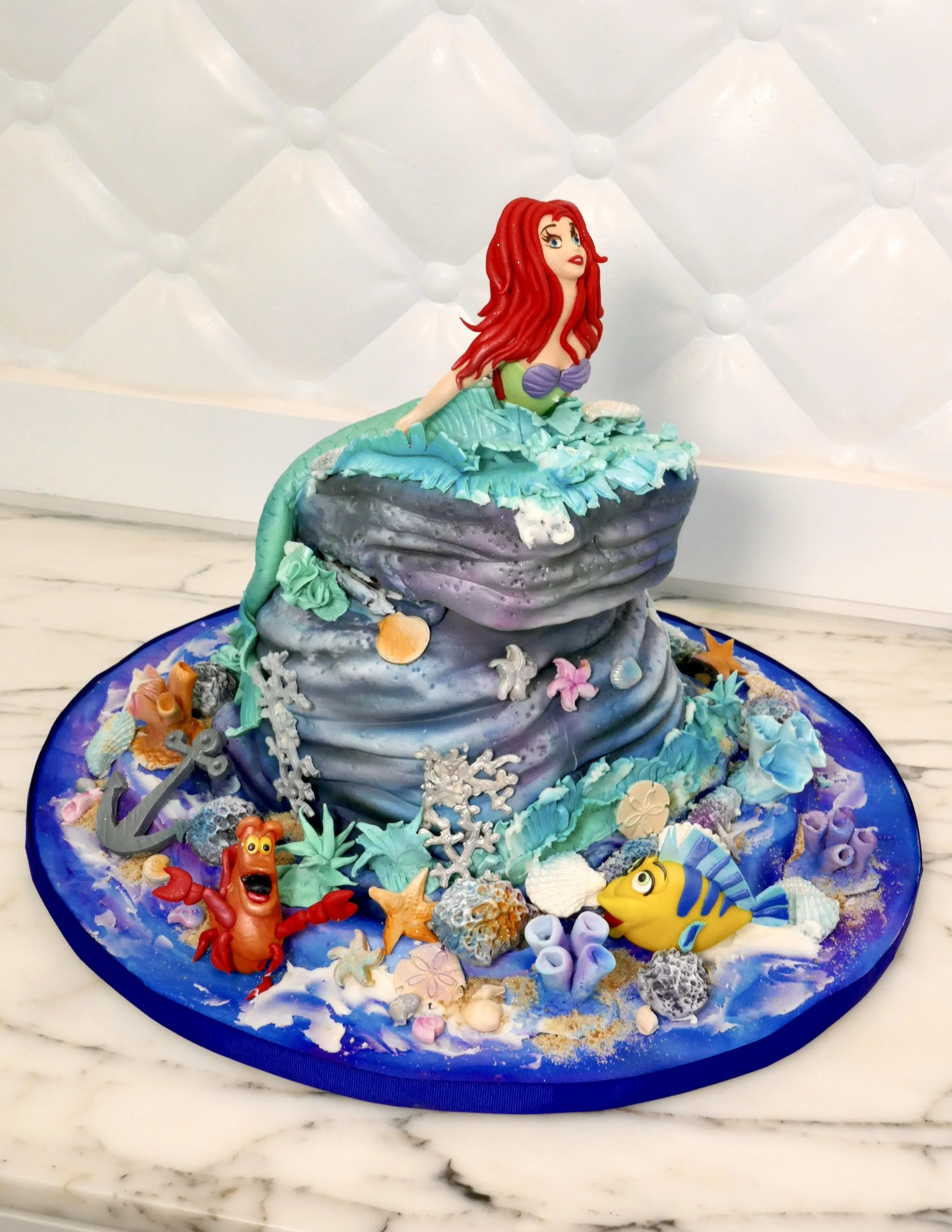 3D under-the-sea themed cake featuring a sculpted mermaid with red hair sitting on ocean waves, surrounded by seashells, coral, starfish, an anchor, and colorful sea creatures on a blue cake board.