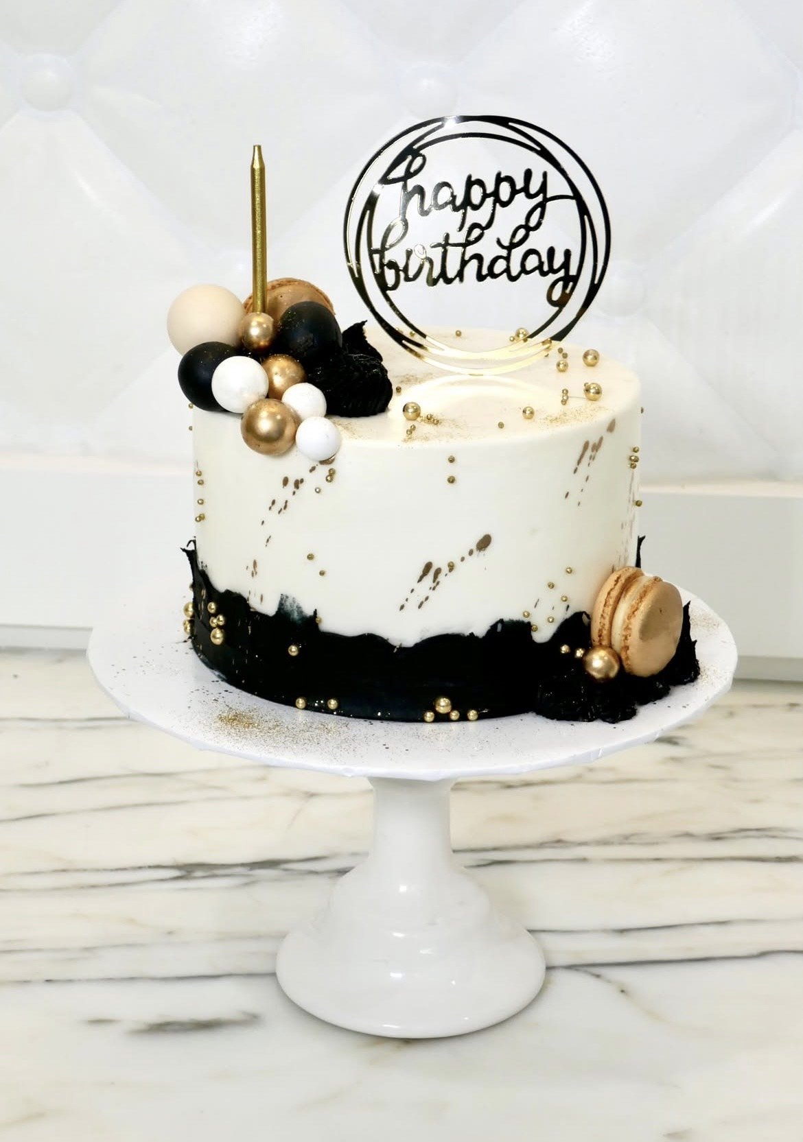 White and black buttercream birthday cake with gold accents, topped with a clear acrylic ‘Happy Birthday’ sign, gold candle, and decorative spheres, displayed on a white cake stand.