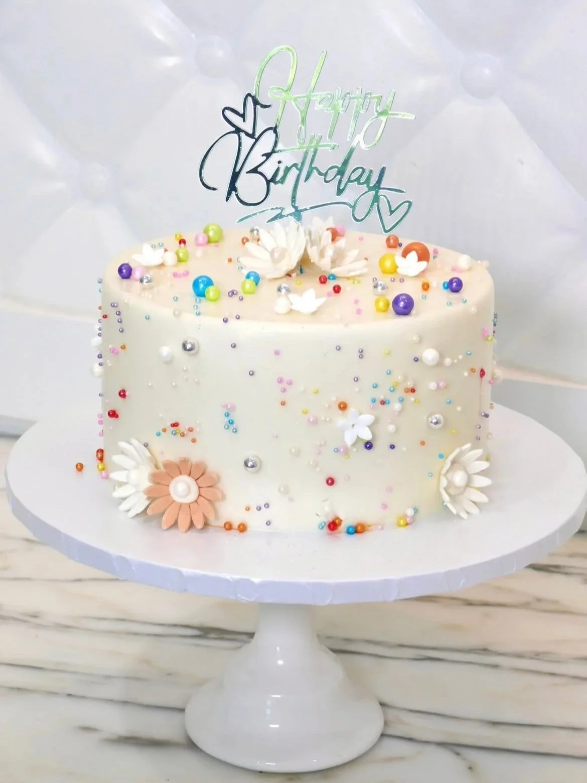 A white buttercream birthday cake decorated with multicolored sprinkles, edible pearls, fondant flowers, and a shiny “Happy Birthday” topper, displayed on a white cake stand.