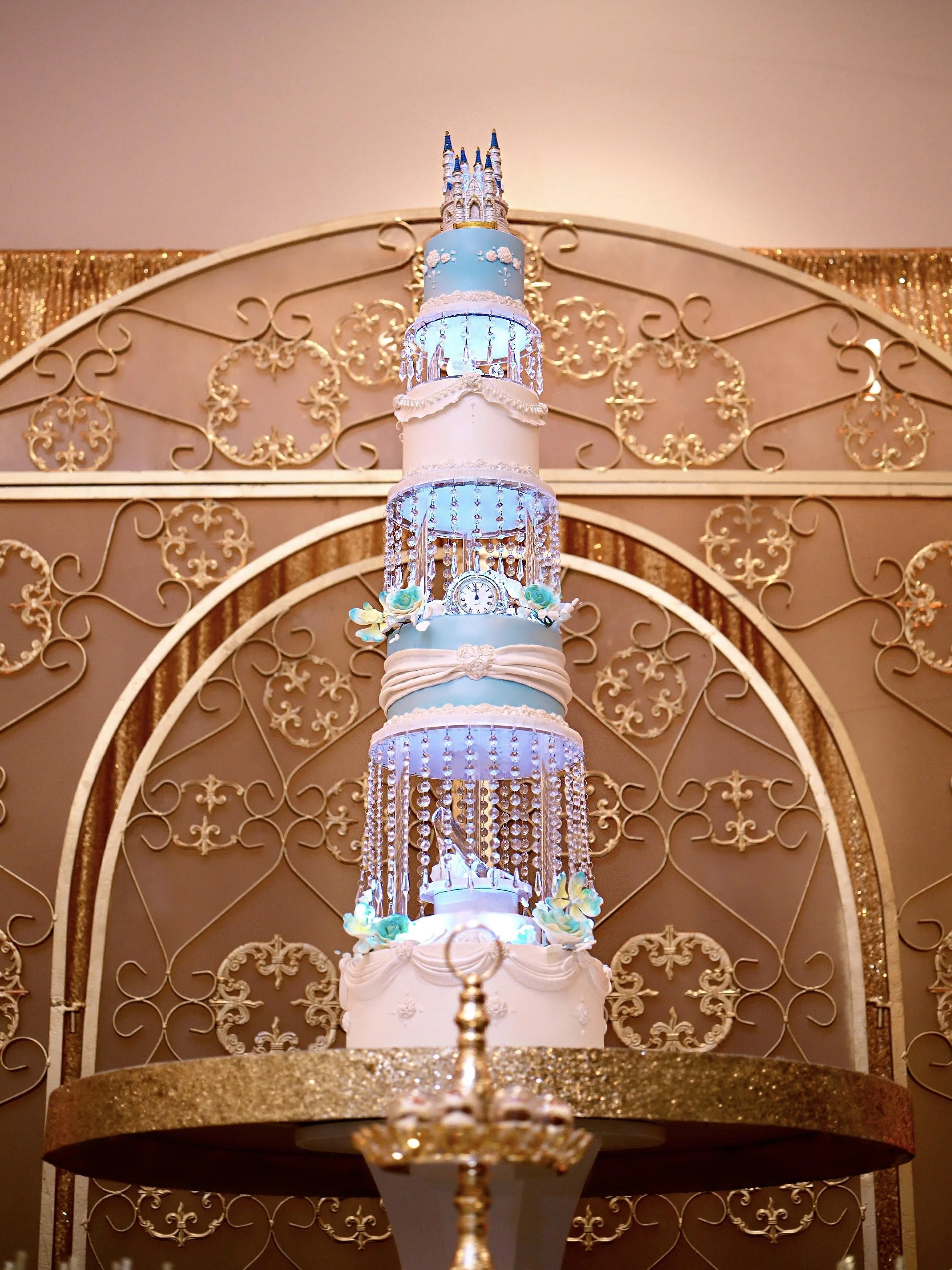 Tall, multi-tier Cinderella-inspired cake in soft blue and white, decorated with crystal strands, floral accents, a clock detail, and a castle topper, displayed against an ornate gold backdrop.