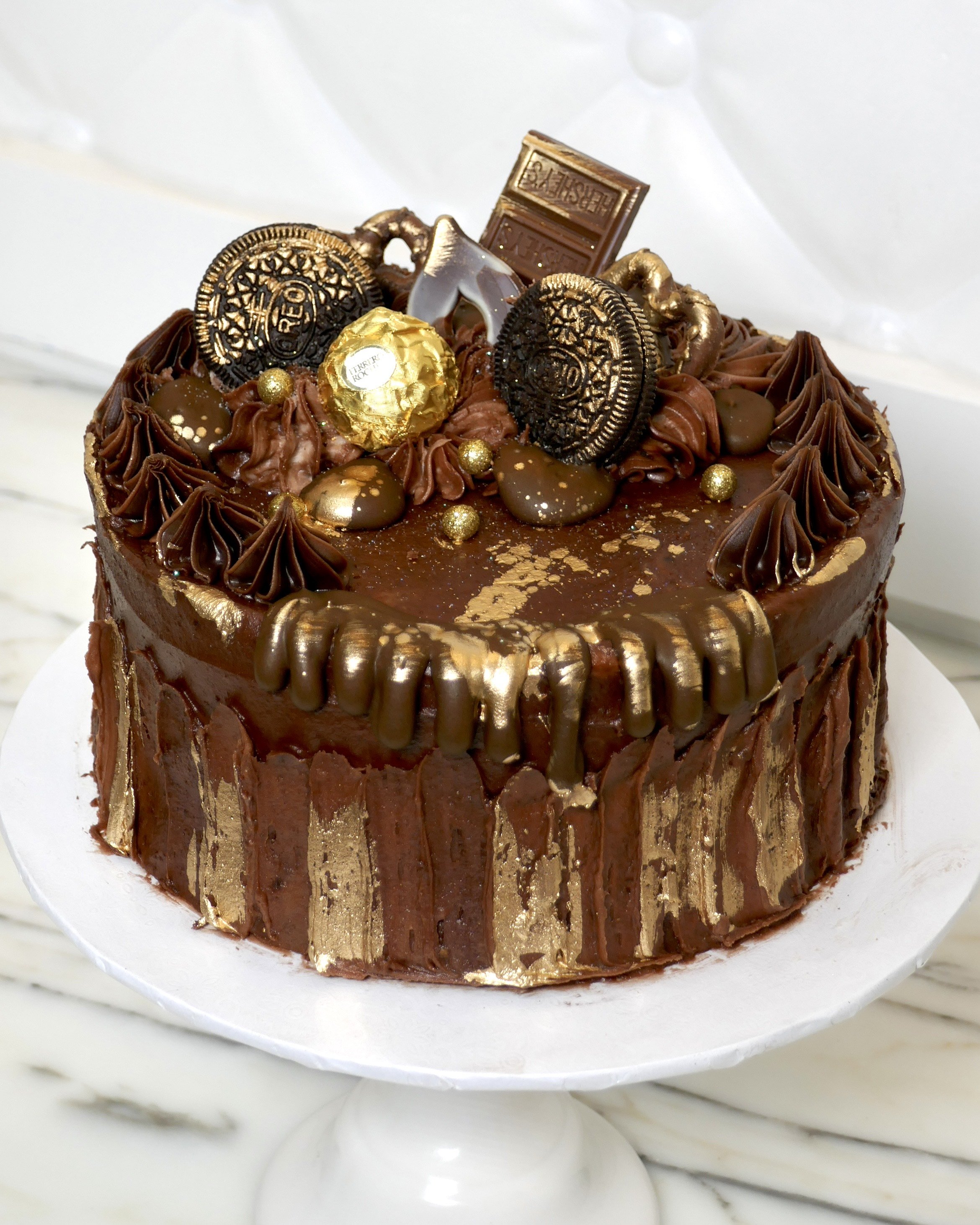 Round chocolate buttercream cake with dark chocolate drip, gold accents, Oreos, Ferrero Rocher, pretzels, and chocolate decorations on top, displayed on a white cake stand.