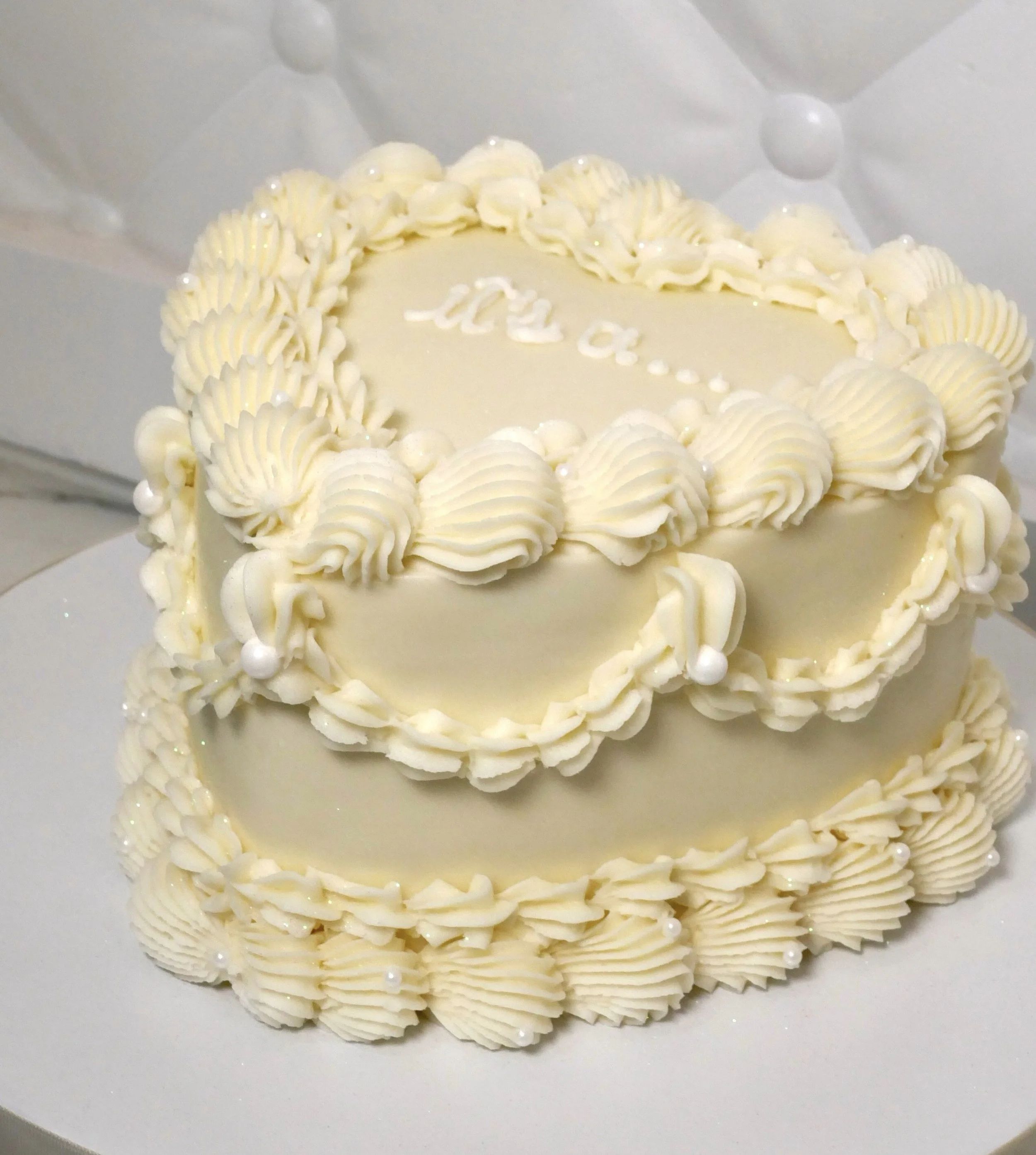 Round vintage-style white buttercream cake decorated with piped shells, swirls, and pearls, that says "It's a..." on top, displayed on a white cake stand over a marble surface