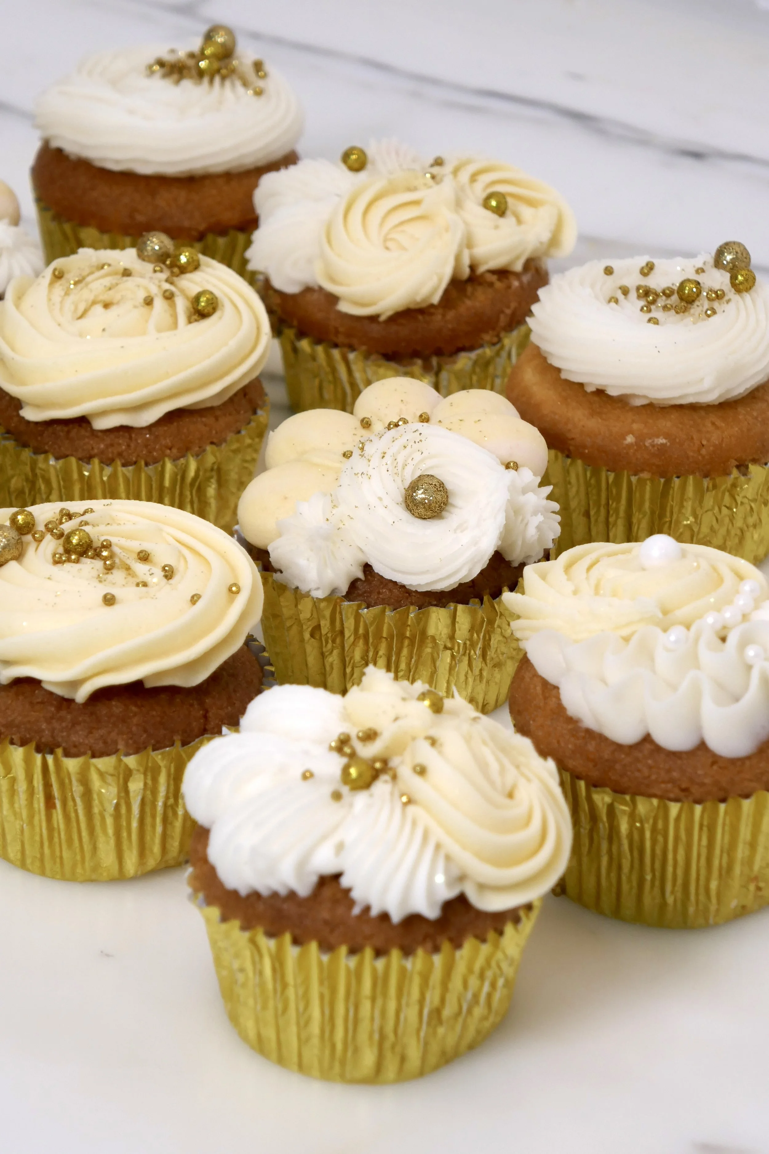 Vanilla cupcakes in gold foil liners topped with swirls of white buttercream and decorated with gold sprinkles and edible pearls, arranged on a white surface.