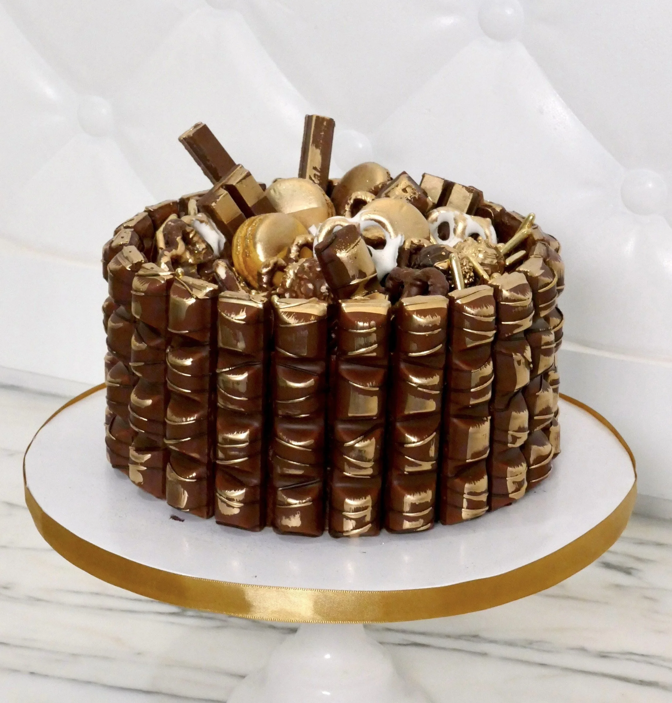 Round chocolate celebration cake wrapped with rows of chocolate bars and topped with gold-dusted macarons, pretzels, and truffles, displayed on a white and gold cake stand.