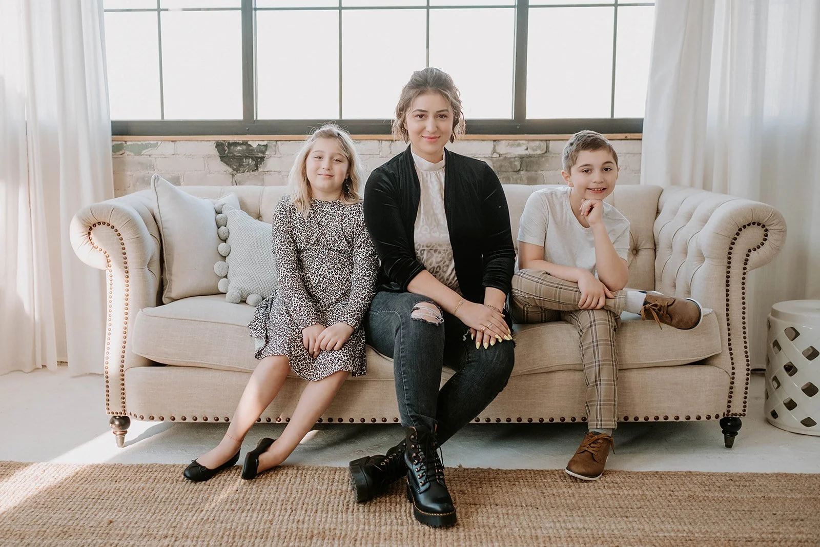 Three people sitting on a beige sofa in a bright room with large windows and white curtains. The person in the middle is wearing a black jacket and black boots, while the others are in casual clothing.