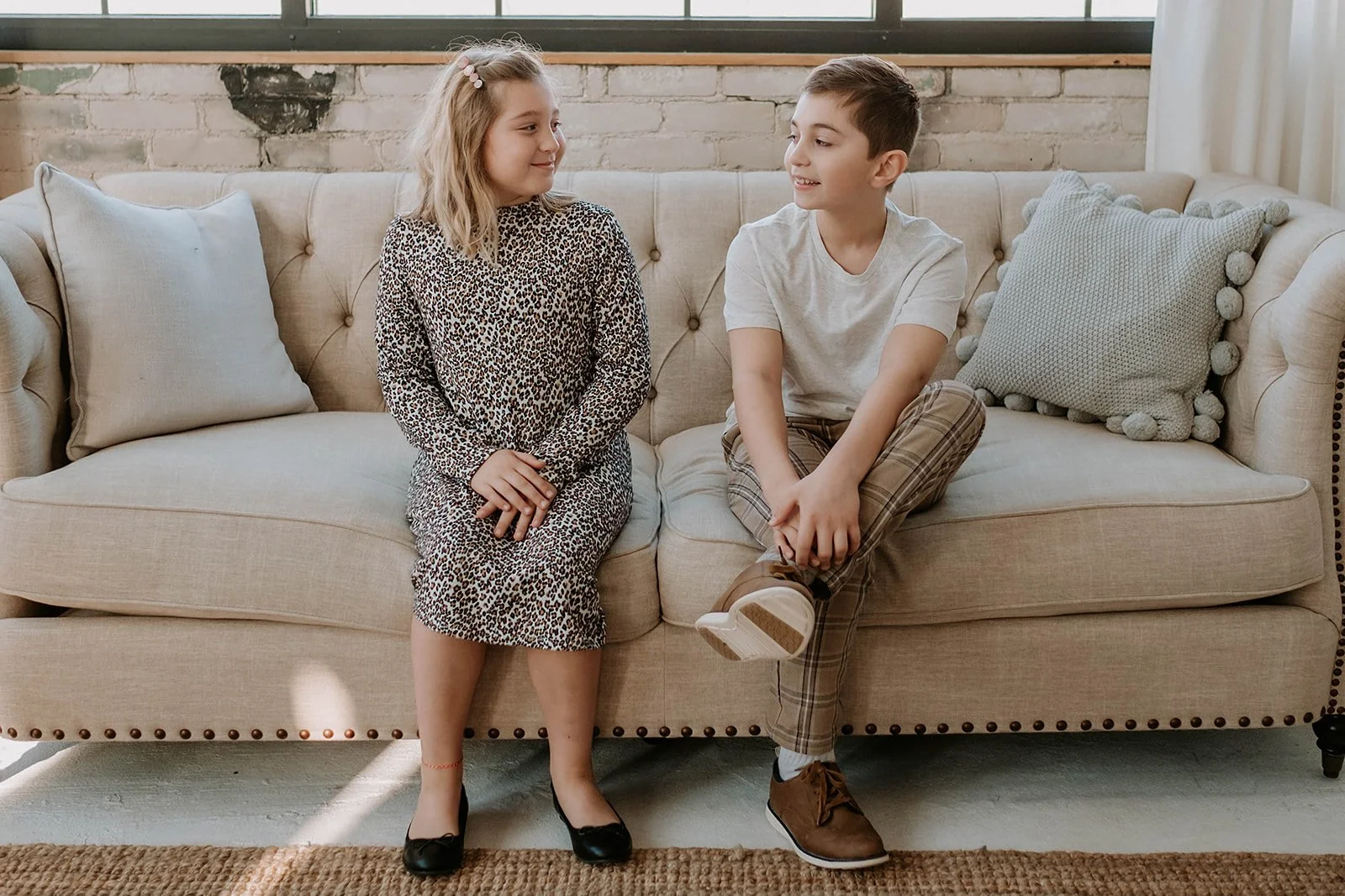 Two children sitting on a beige sofa, a girl in a leopard print dress and a boy in a gray t-shirt with plaid pants, smiling at each other.
