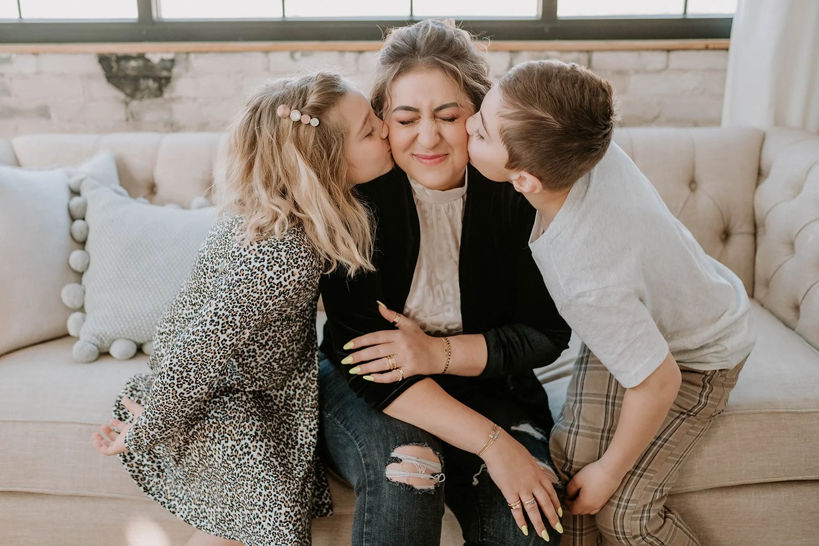 Two children kissing an adult on the cheeks, sitting on a beige couch with cushions in a well-lit room.