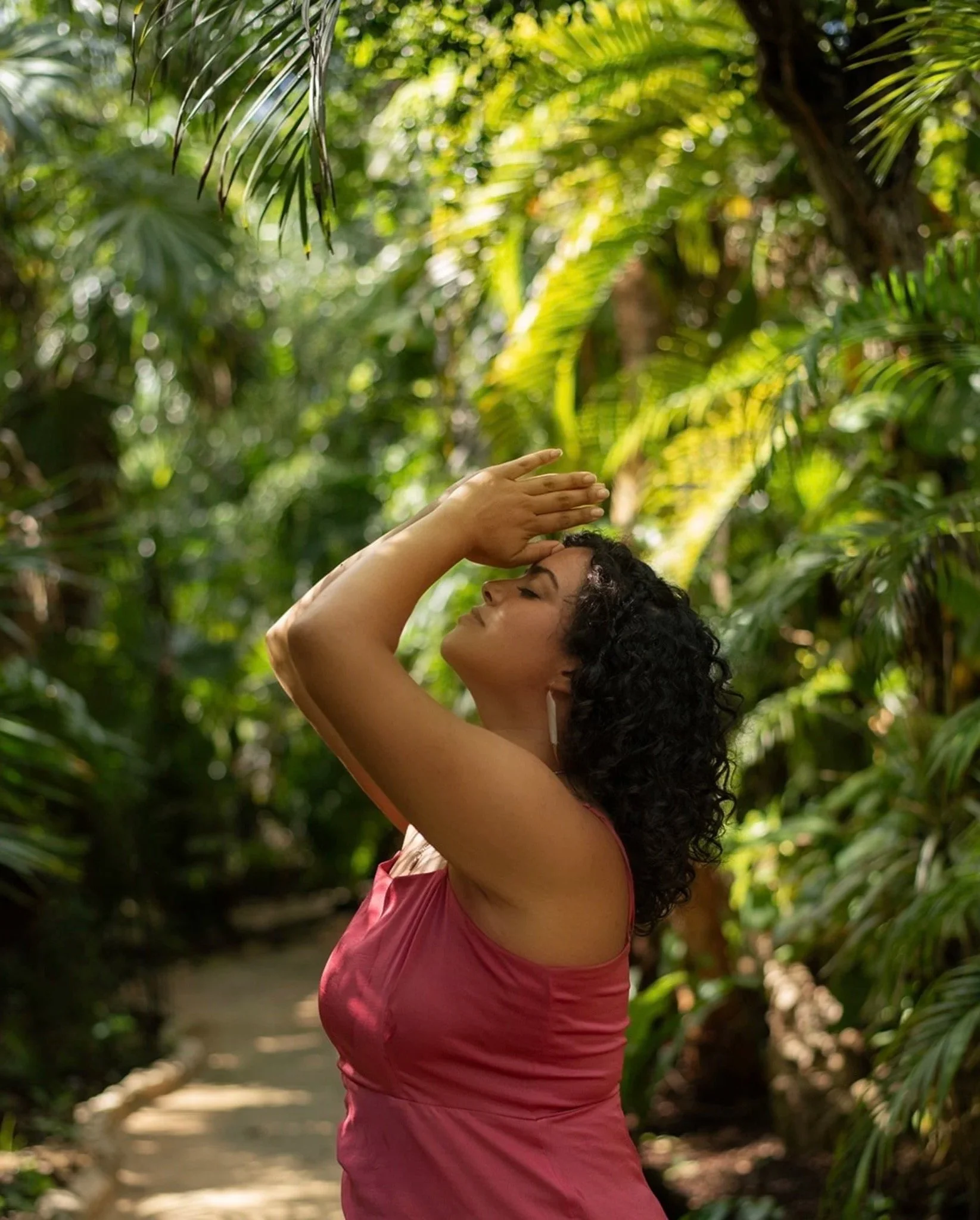 A woman with curly hair wearing a sleeveless pink top standing among lush green foliage with sunlight filtering through.