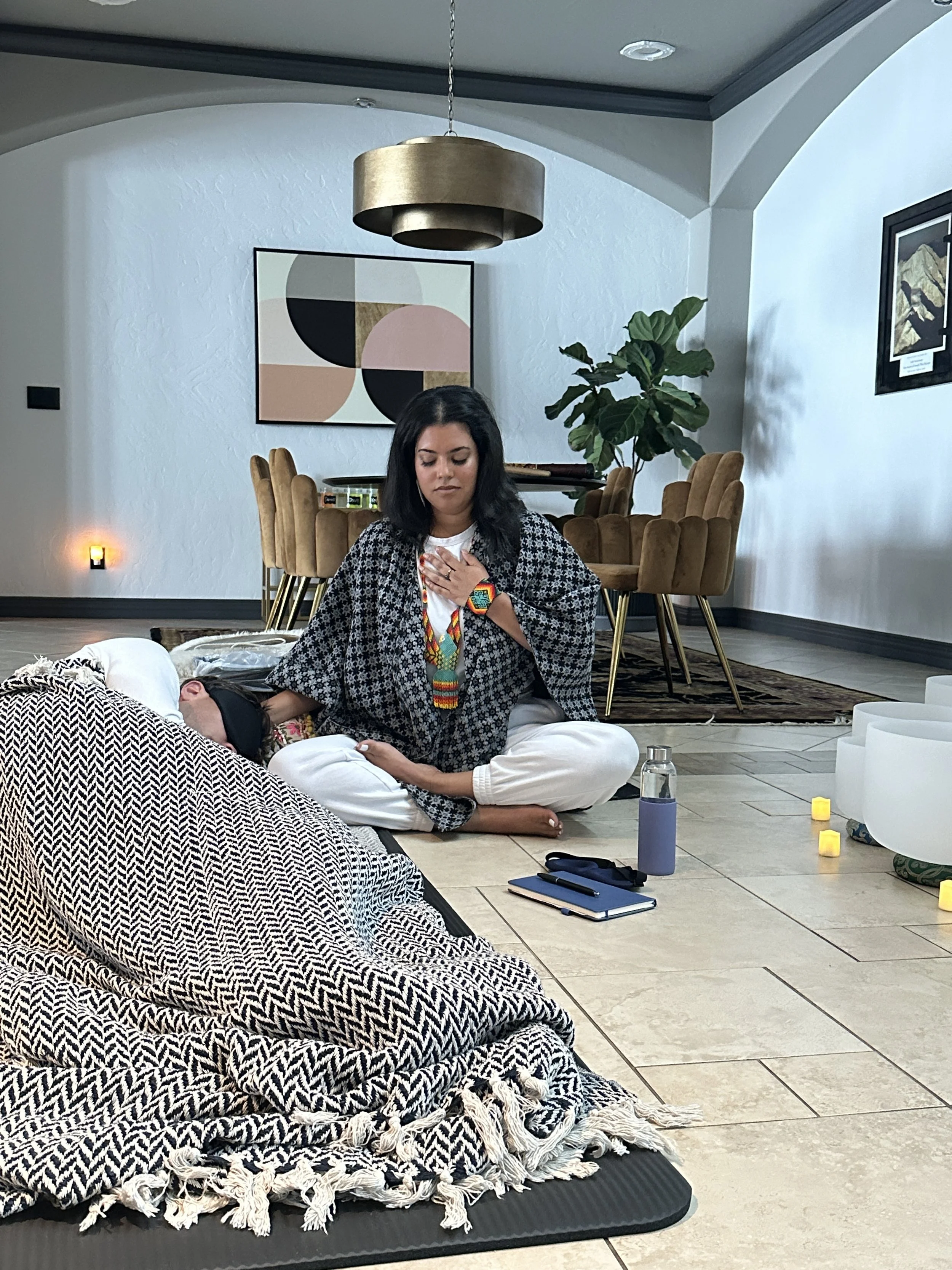 A woman sitting cross-legged on the floor with her eyes closed and hand on her chest, appearing to meditate or pray, in a modern living room with abstract wall art and dining table in the background.