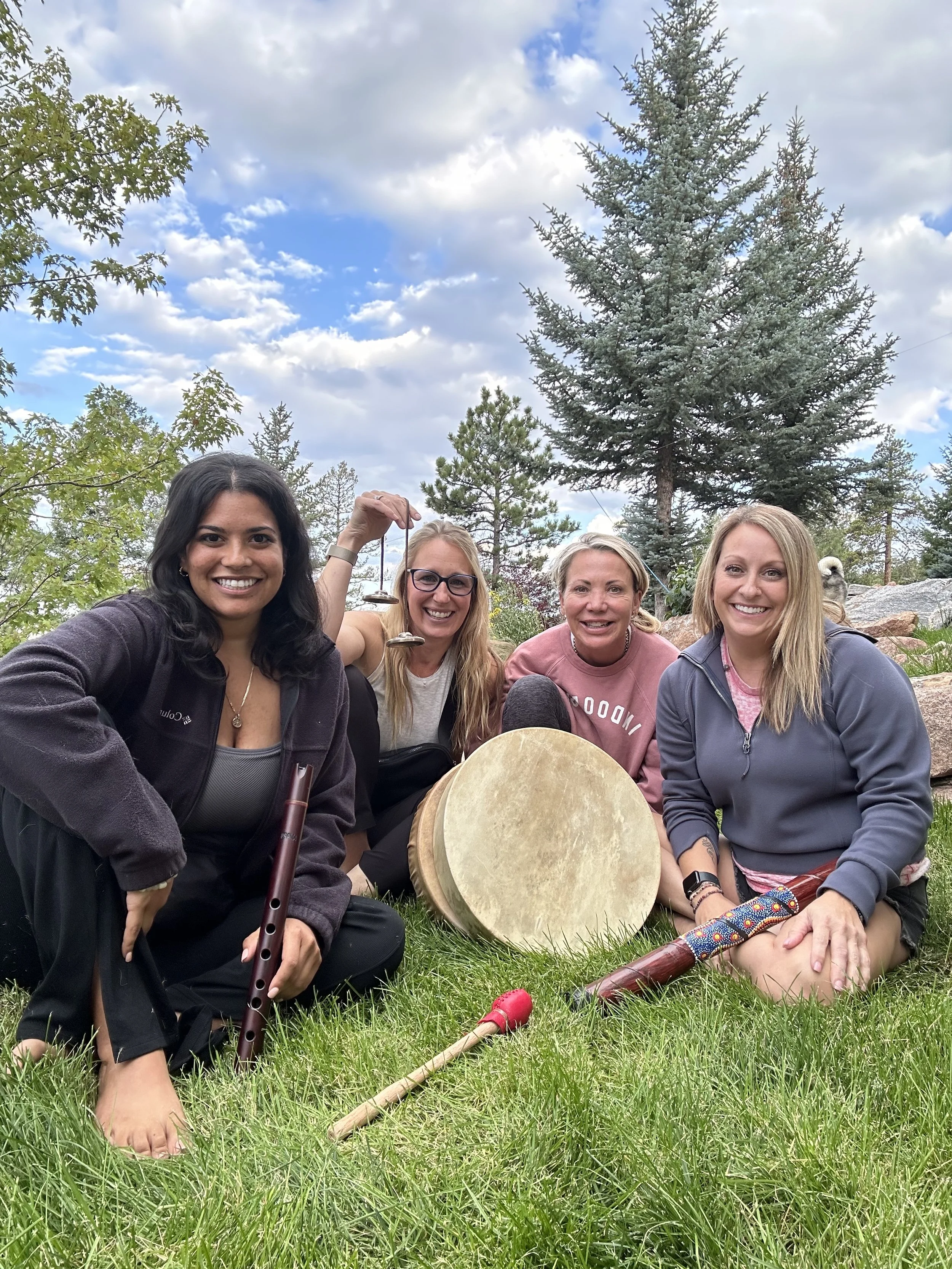 Group of four women sitting on grass outdoors, smiling, with musical instruments including a flute and a drum, in front of trees and a partly cloudy sky.