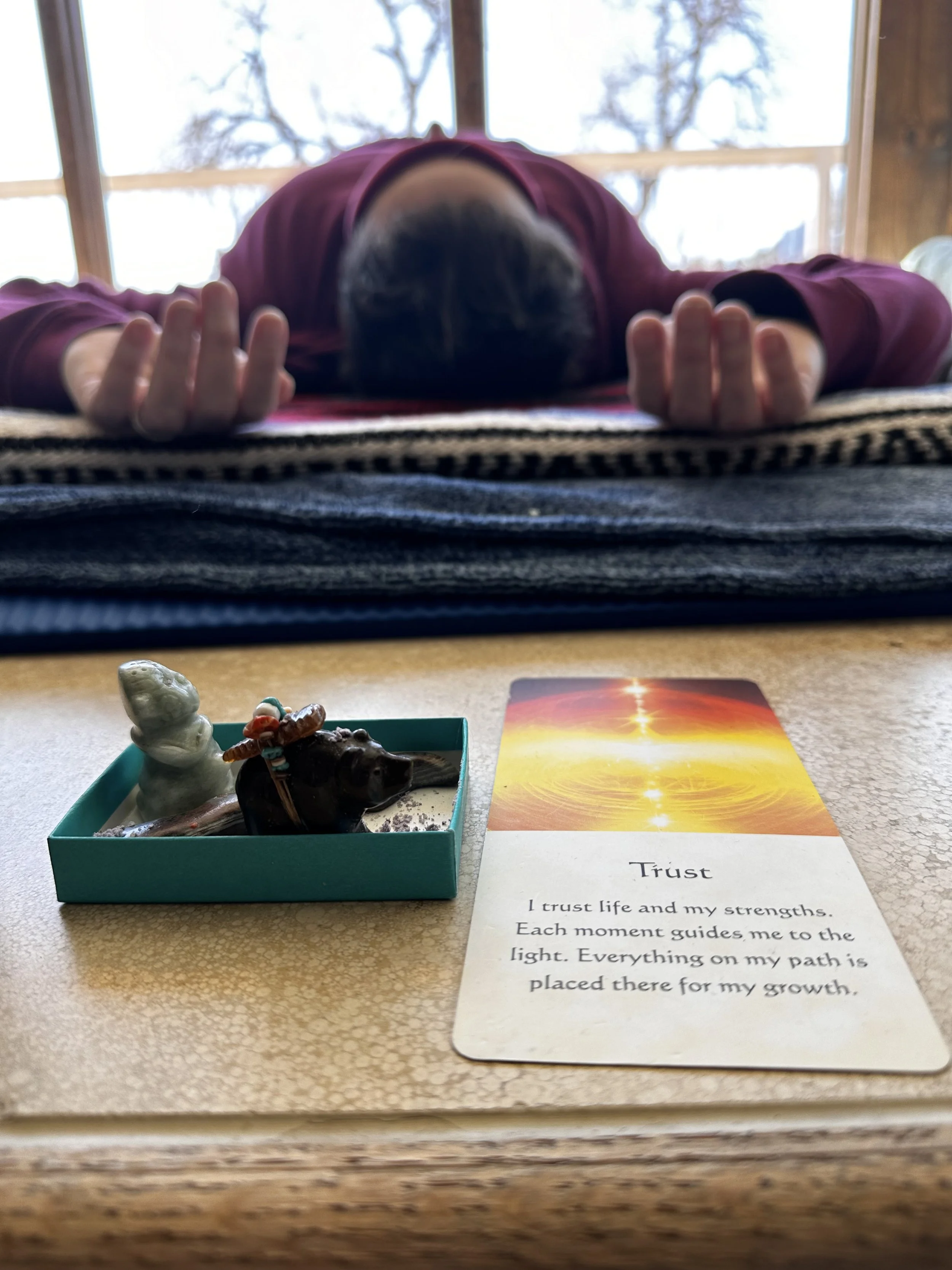 A close-up of a wooden table holding a small teal box with a white figurine of a unicorn and a miniature black buffalo with a painted buffalo horn and bead necklace. Next to the box is a card with the word 'Trust' and an inspirational message about trust, life, and growth. In the background, a person is lying face down on a woven mat with their arms extended, inside a room with large windows showing trees outside.