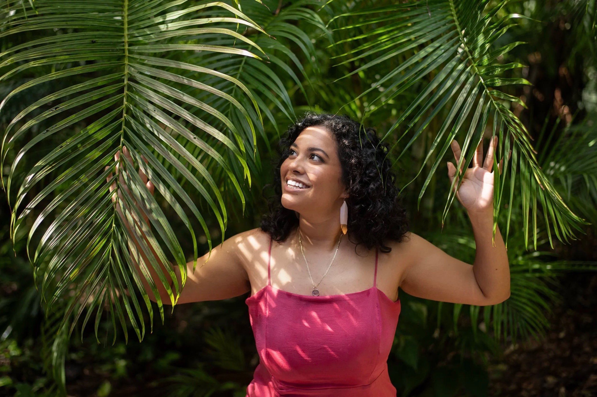 Woman with dark curly hair wearing a pink strapless dress standing among green tropical palm leaves.