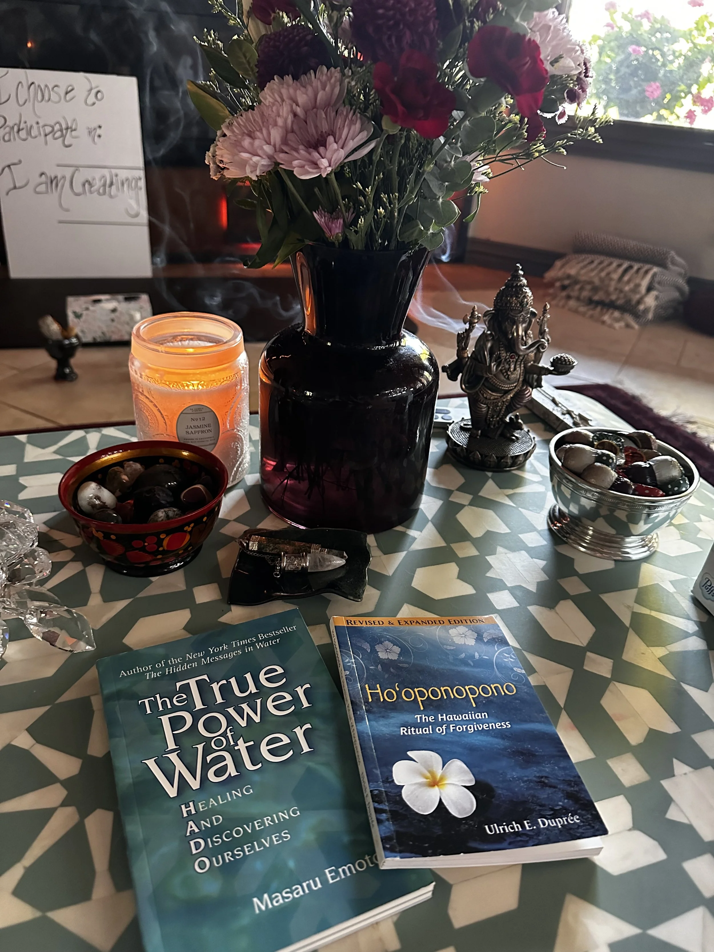 A table with a bouquet of pink, red, and white flowers in a black vase, a lit candle, a silver figurine of Lord Ganesha, bowls with stones or crystals, and two books titled 'The True Power of Water' by Masaru Emoto and 'Ho'oponopono' by Ulrich E. Dupré. A handwritten sign is visible in the background.